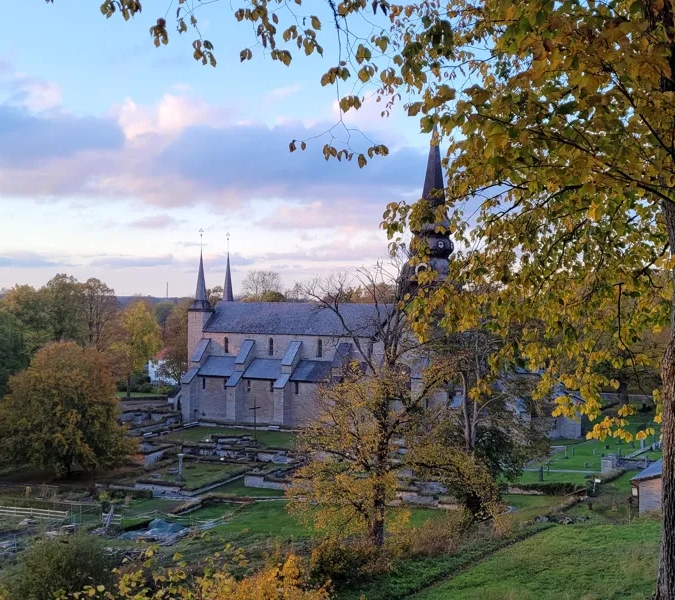 En bild på Varnhems klosterkyrka, en stor kyrka i ljus sten, inbäddad i höstfärger.
