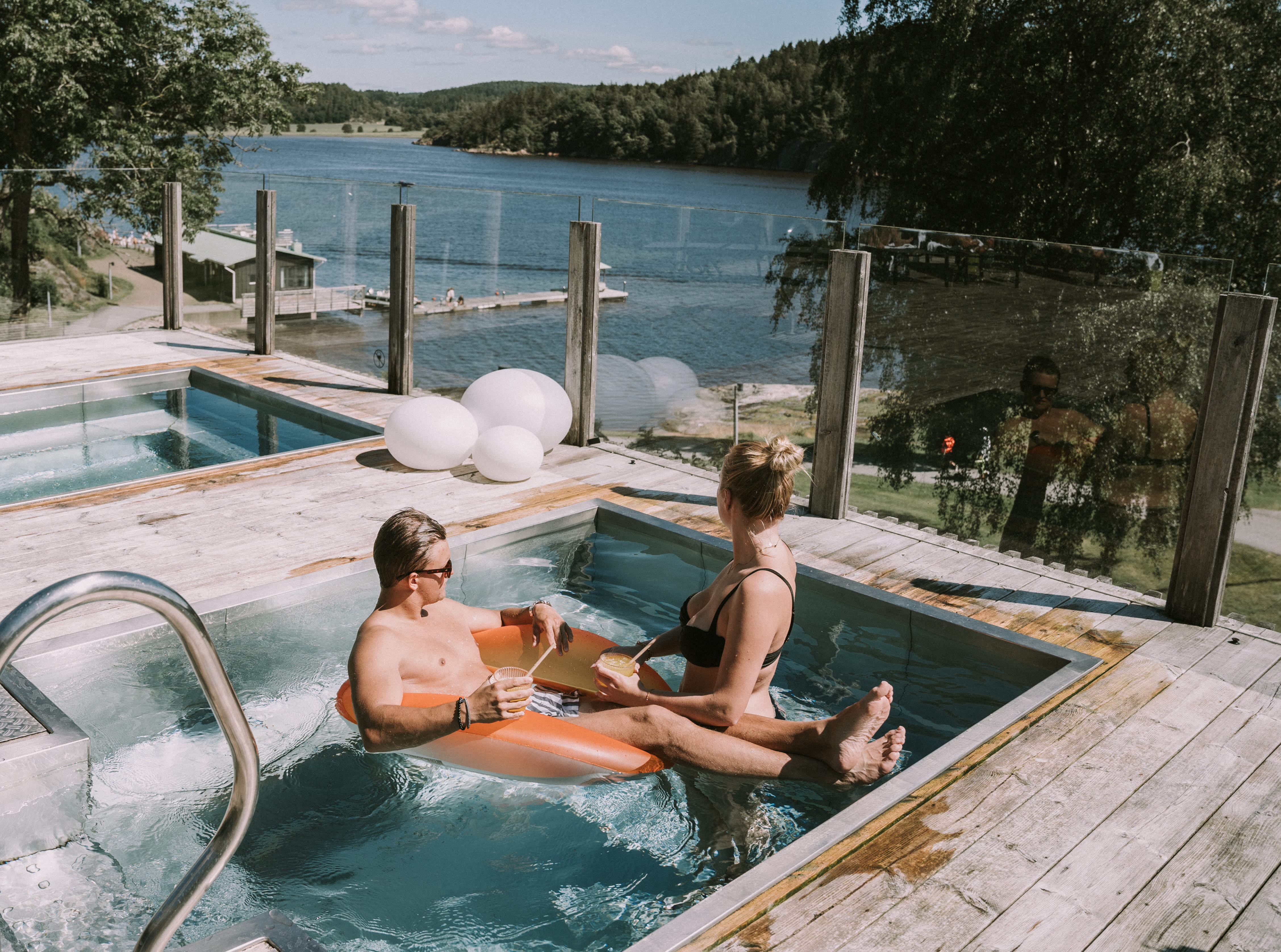 Man and woman, hotel guests in the outdoor pool in summertime, overlooking the Gullmarsfjorden with drinks in their hands
