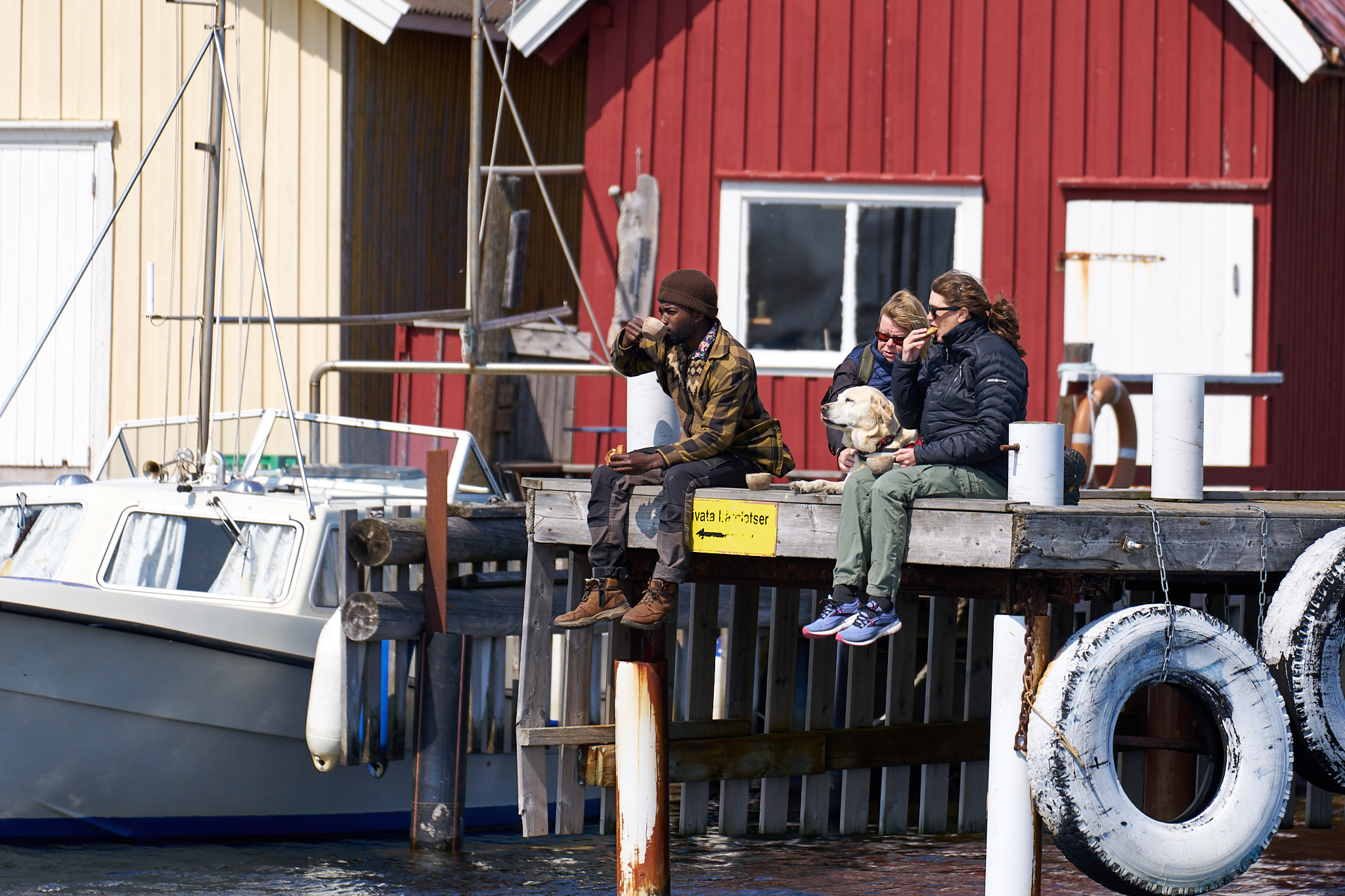 Three people have a fika on a dock in Brevik, in front of boathouses and a boat, in Kosterhavet National Park