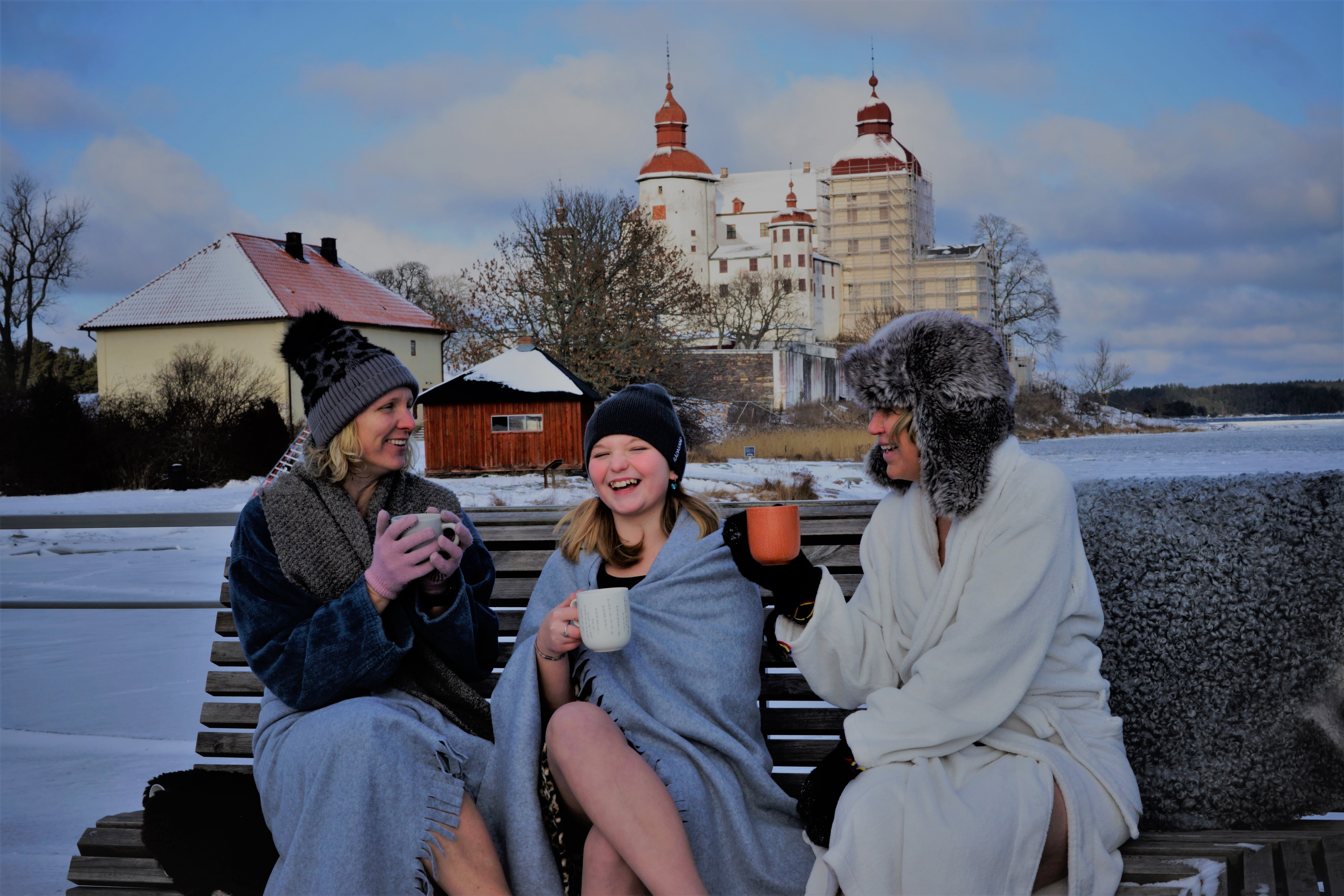Three women winter bathing sit on a bench and drink something warming by Läckö Castle