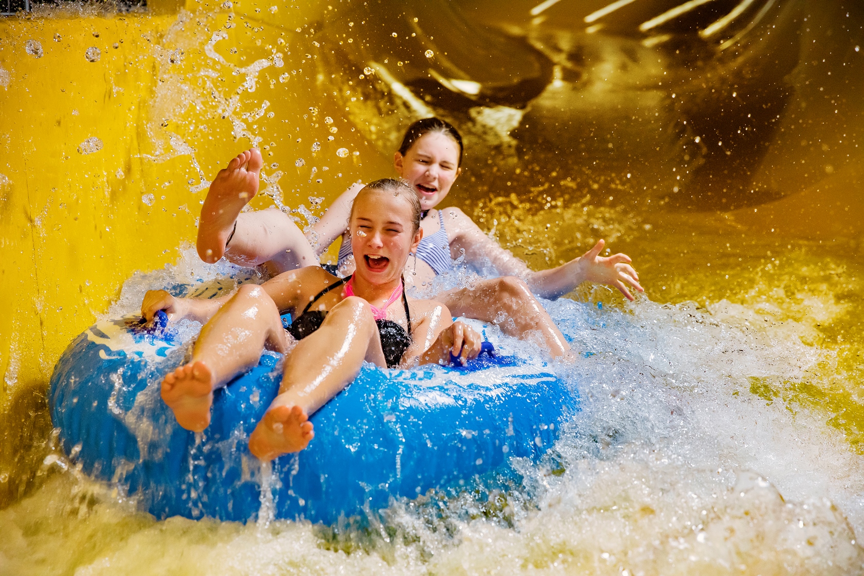 Two girls are laughing and riding a yellow water slide with water splashing around.