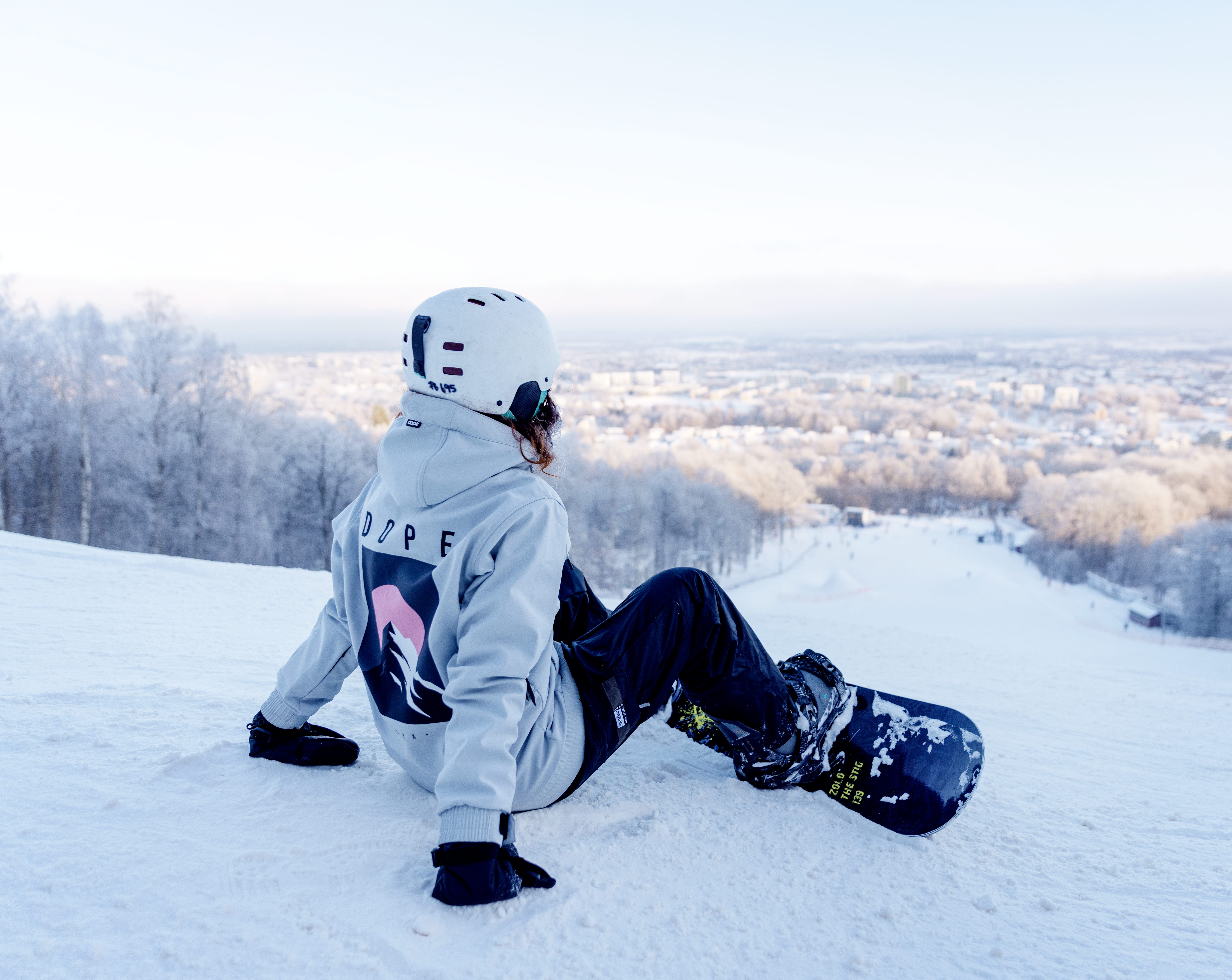 Person with helmet and snowboard sits in Billingebacken and looks out over the hill and the town of Skövde
