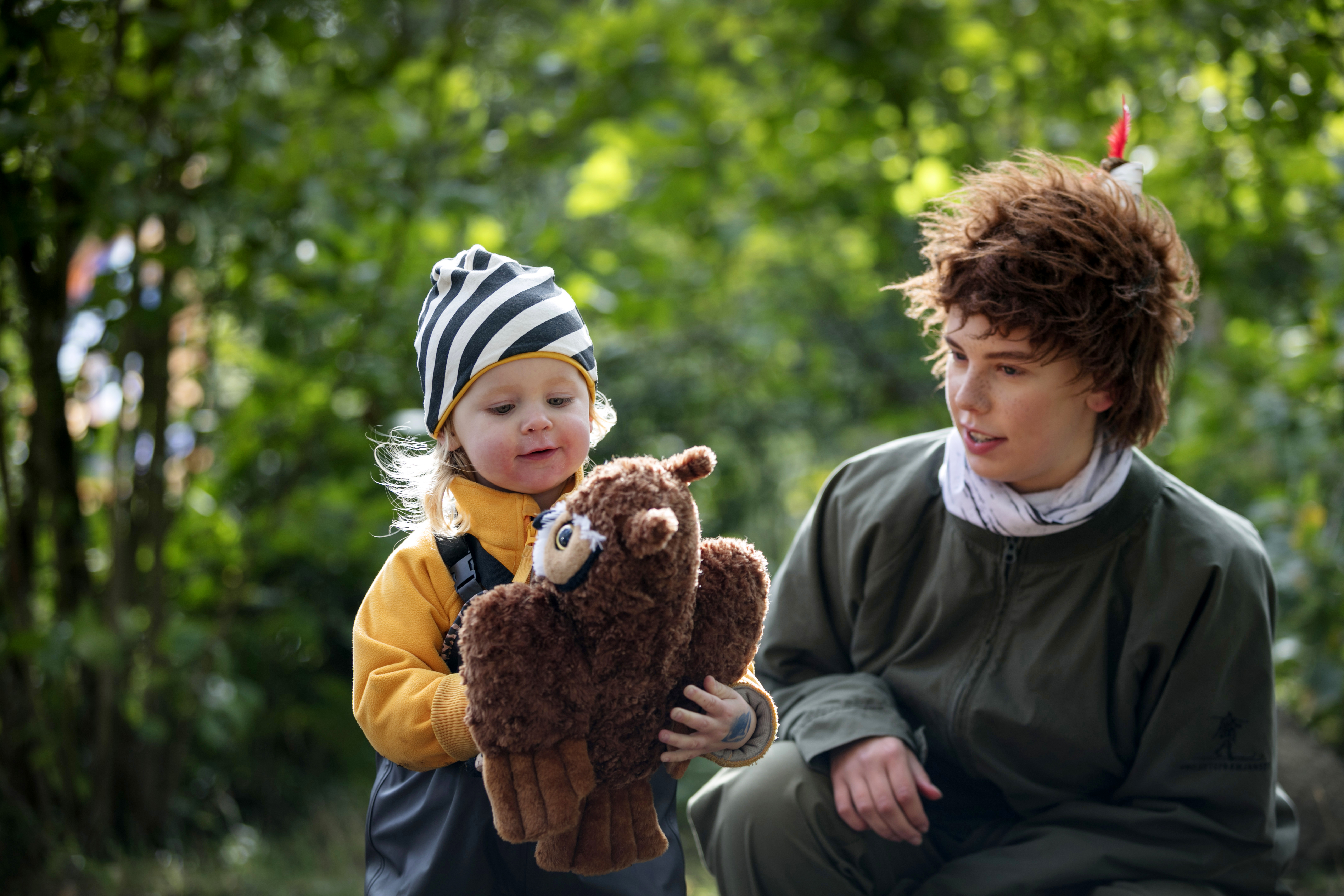 Girl in yellow sweater and black / white striped hat plays with a teddy bear together with Skogsmulle.