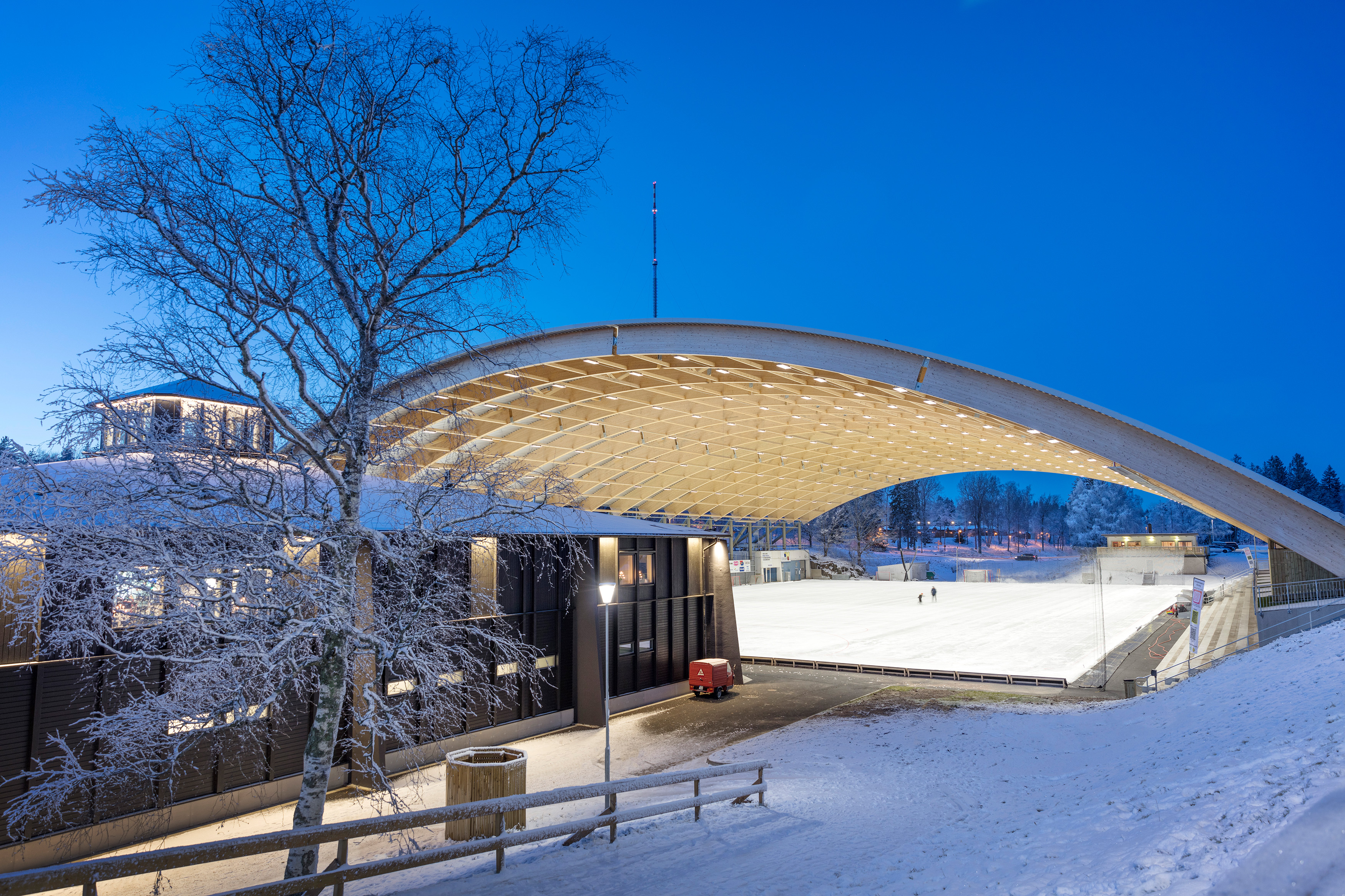 Illuminated black building (Billingecenter) at dusk. There is snow on the slopes and in the background you can see the vaulted roof over the rink.