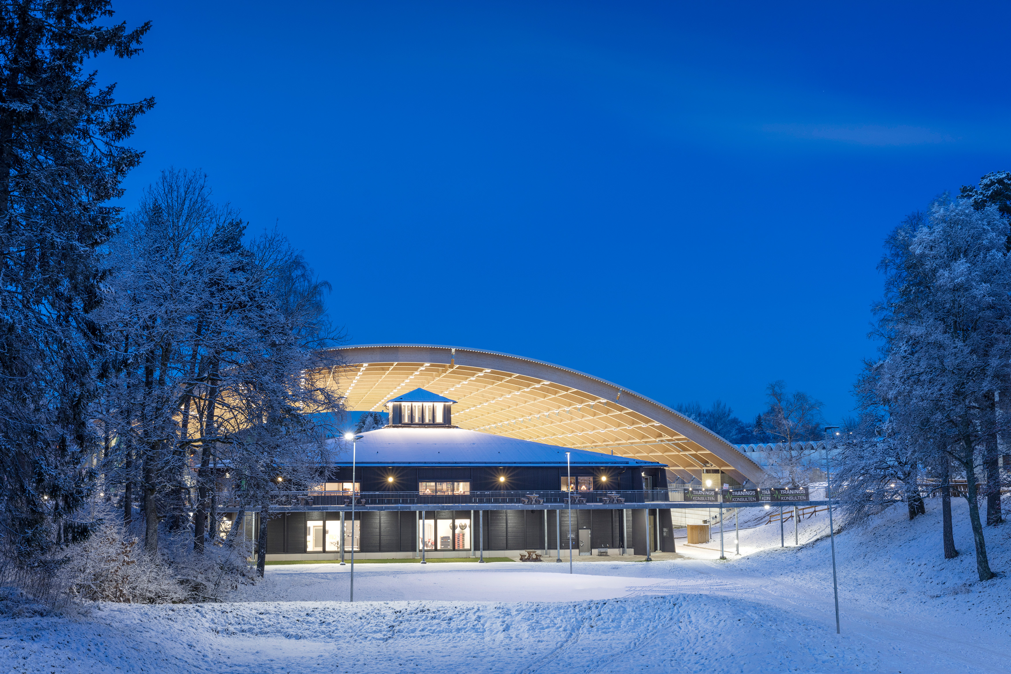 Illuminated black building (Billingecenter) at dusk. There is snow on the slopes and in the background you can see the vaulted roof over the rink.