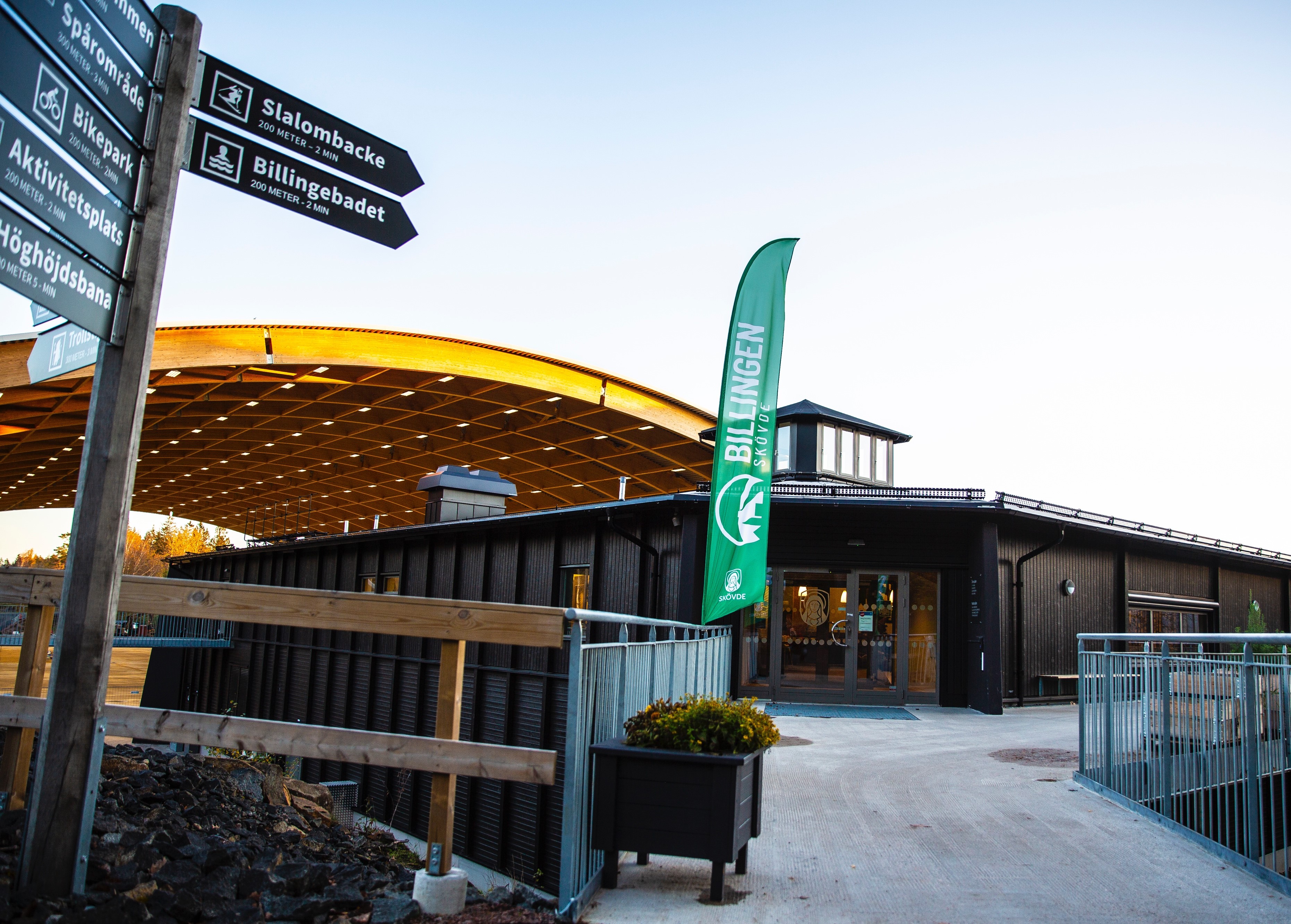 The footbridge to a low black wooden building with a glass dome on the roof - Billingecenter. To the left is a sign with several color indicators for exercise tracks, bike tracks etc. In the background, the vaulted roof over the rink can be seen.