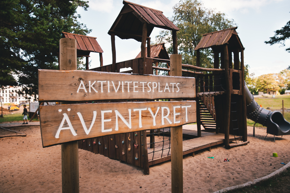 Wooden sign with the text "Activity site THE ADVENTURE". Behind it, you can see an all-wooden playground on two floors, a slide, surrounded by sand.