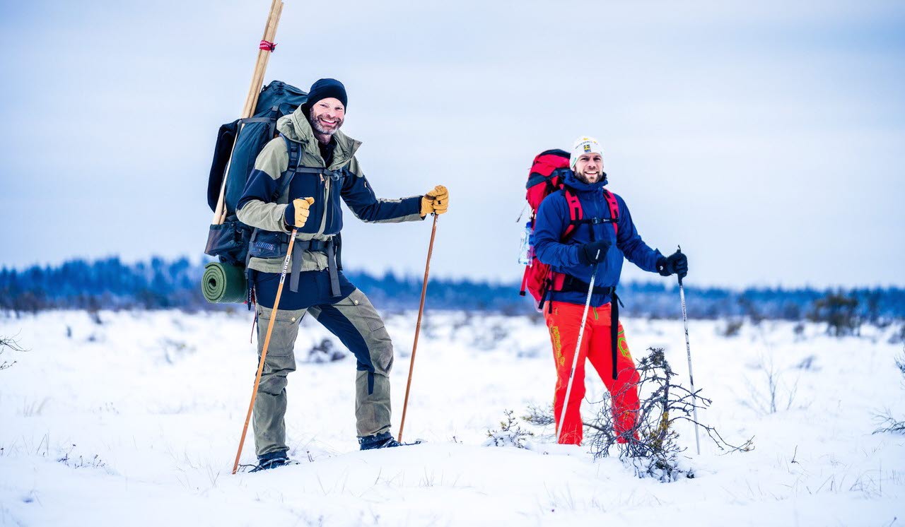 Two men are standing in the middle of a winter landscape with full hiking gear and snowshoes on their feet.