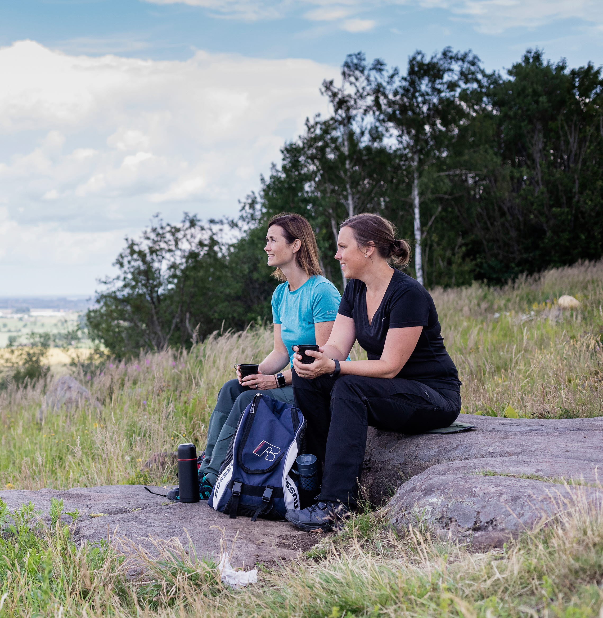 Two women sitting on a rock and having coffee and looking at the view.