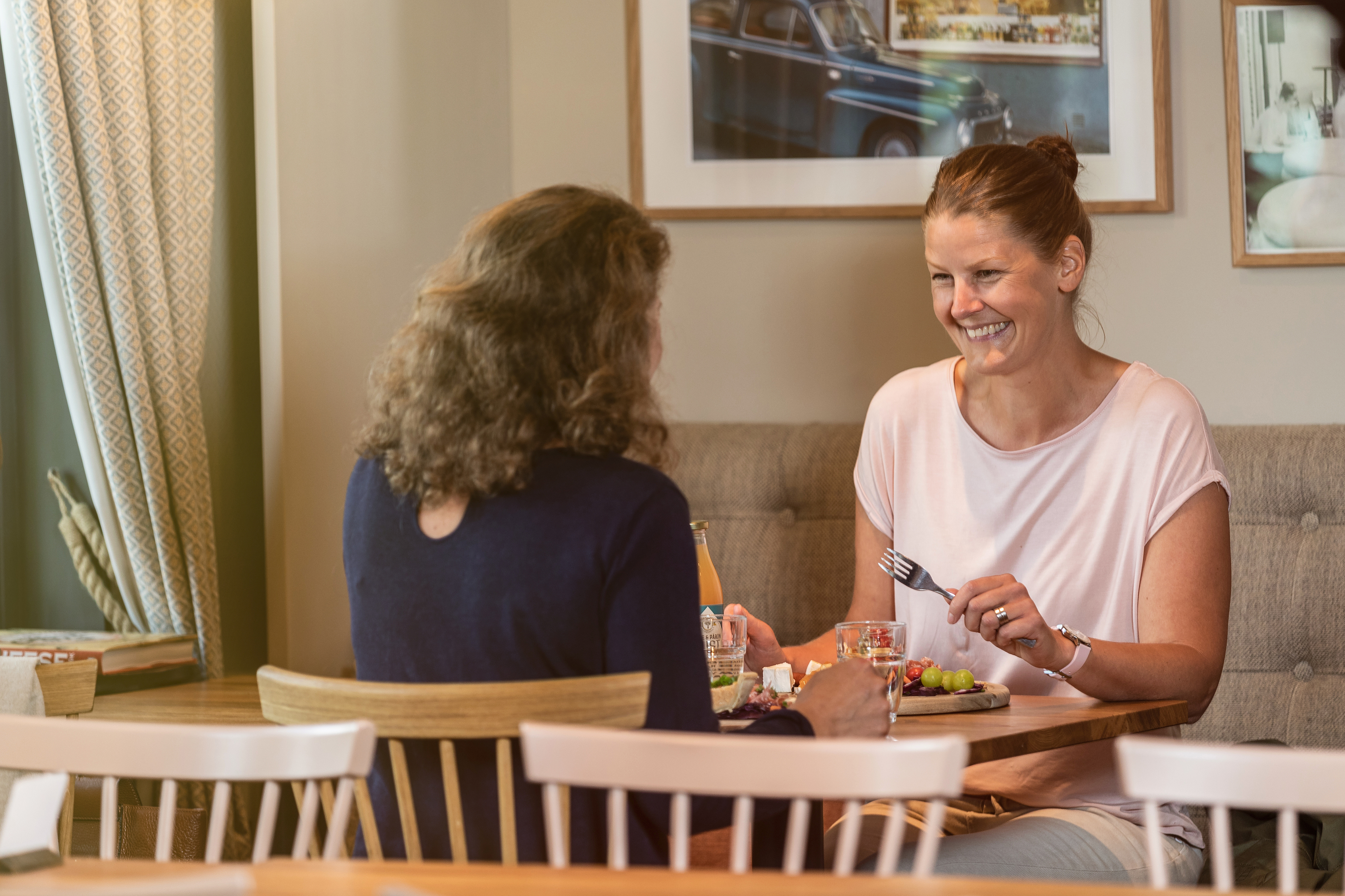 Two women sitting and eating cheese platter and laughing and talking.