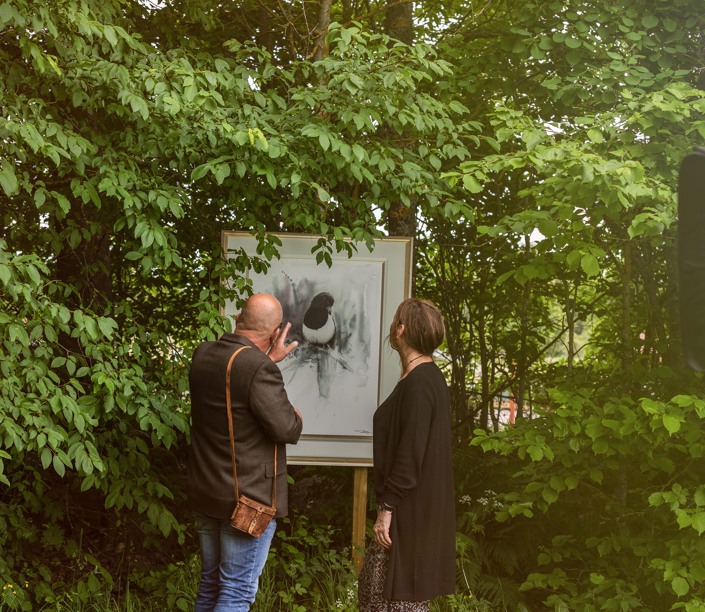 Man and woman peeking at a painting outdoors among lush leaves.
