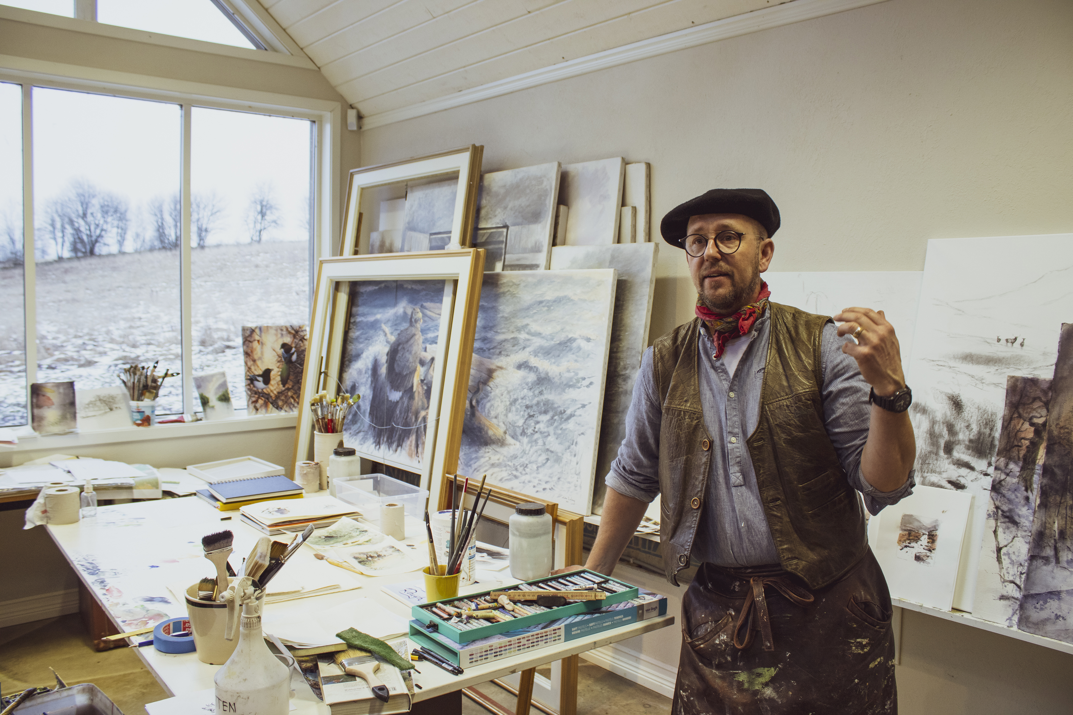 A man with a beret and an apron in leather stands in front of his painted paintings. Brushes on a table next to it.
