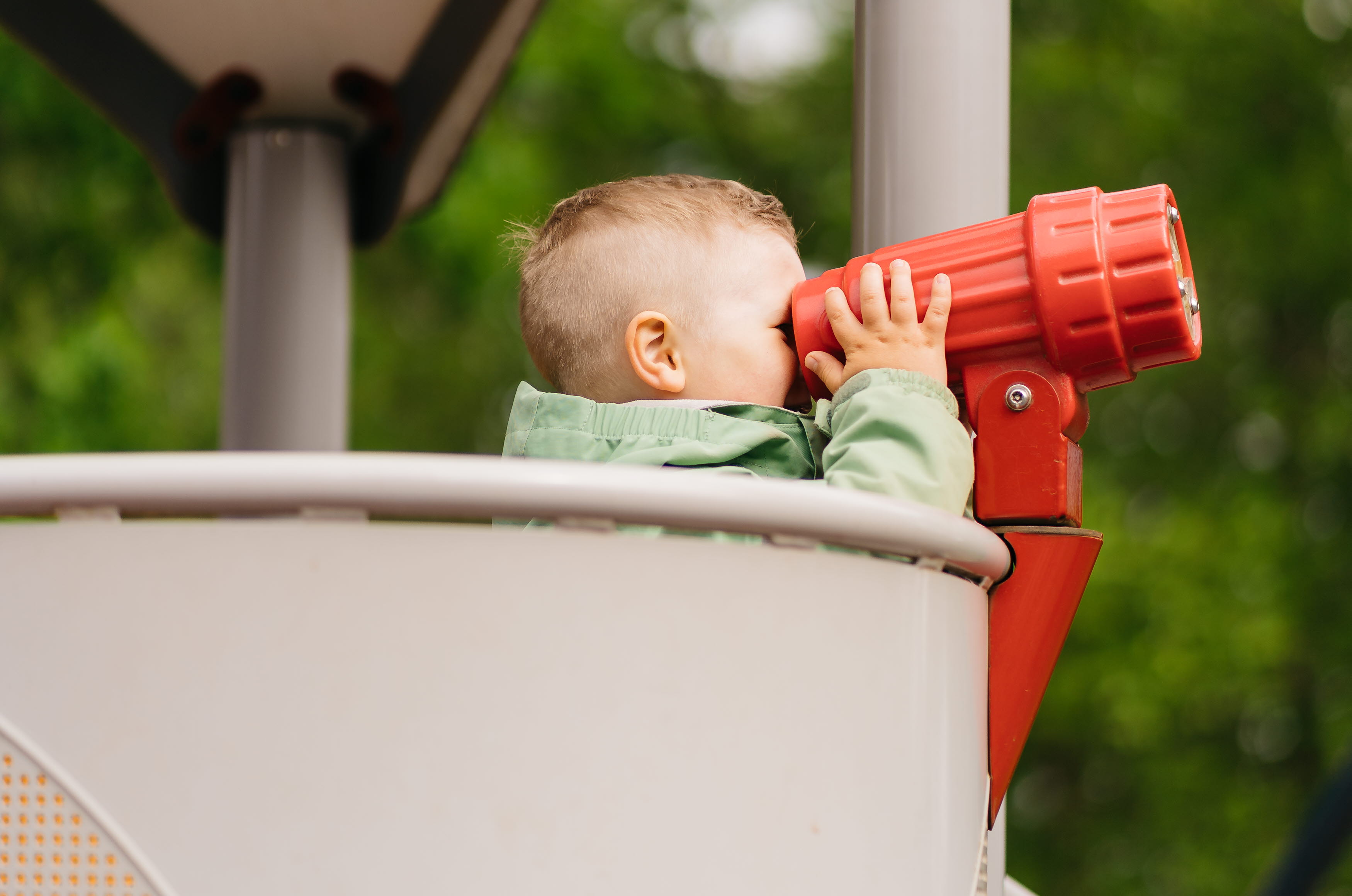 Boy standing in a playground and peeking in a red plastic binoculars.