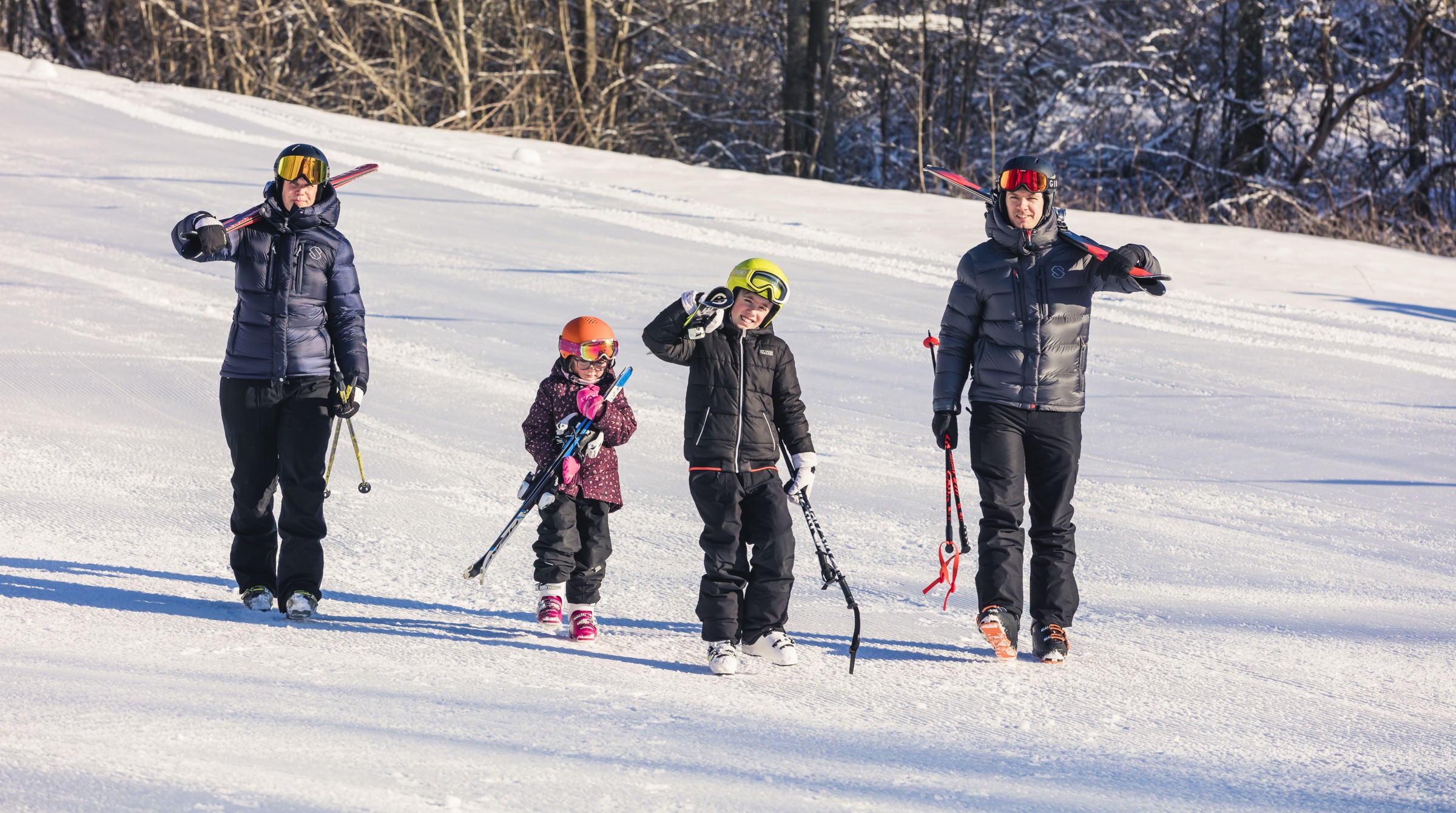A family with two adults and two children walks in the snow and carries their skis.