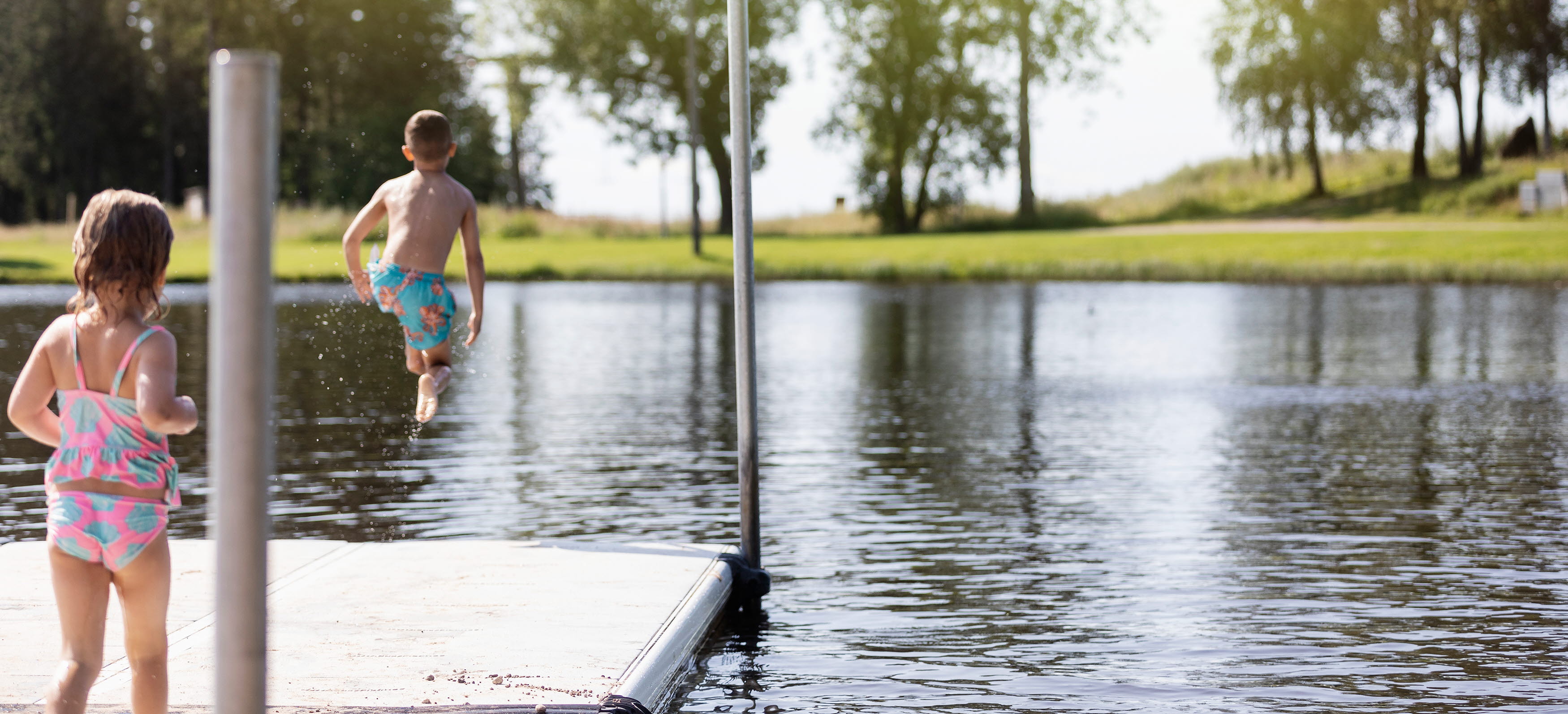 Two children on a jetty with their backs to the camera. One of them jumps into the water.
