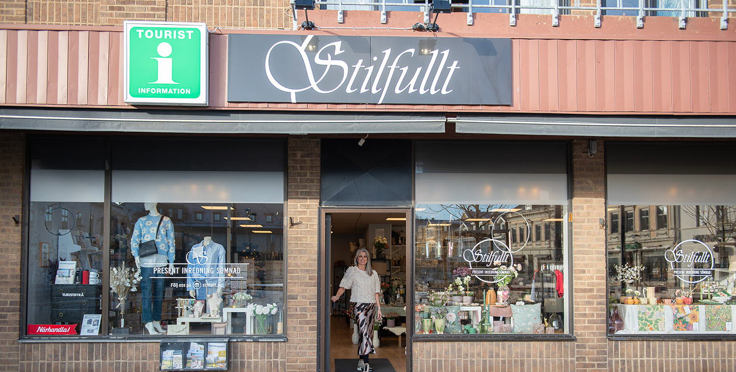 Exterior view of the Tourist Information Center. Large display windows in a brick building. In the open doorway stands Sara, who runs the tourist center.