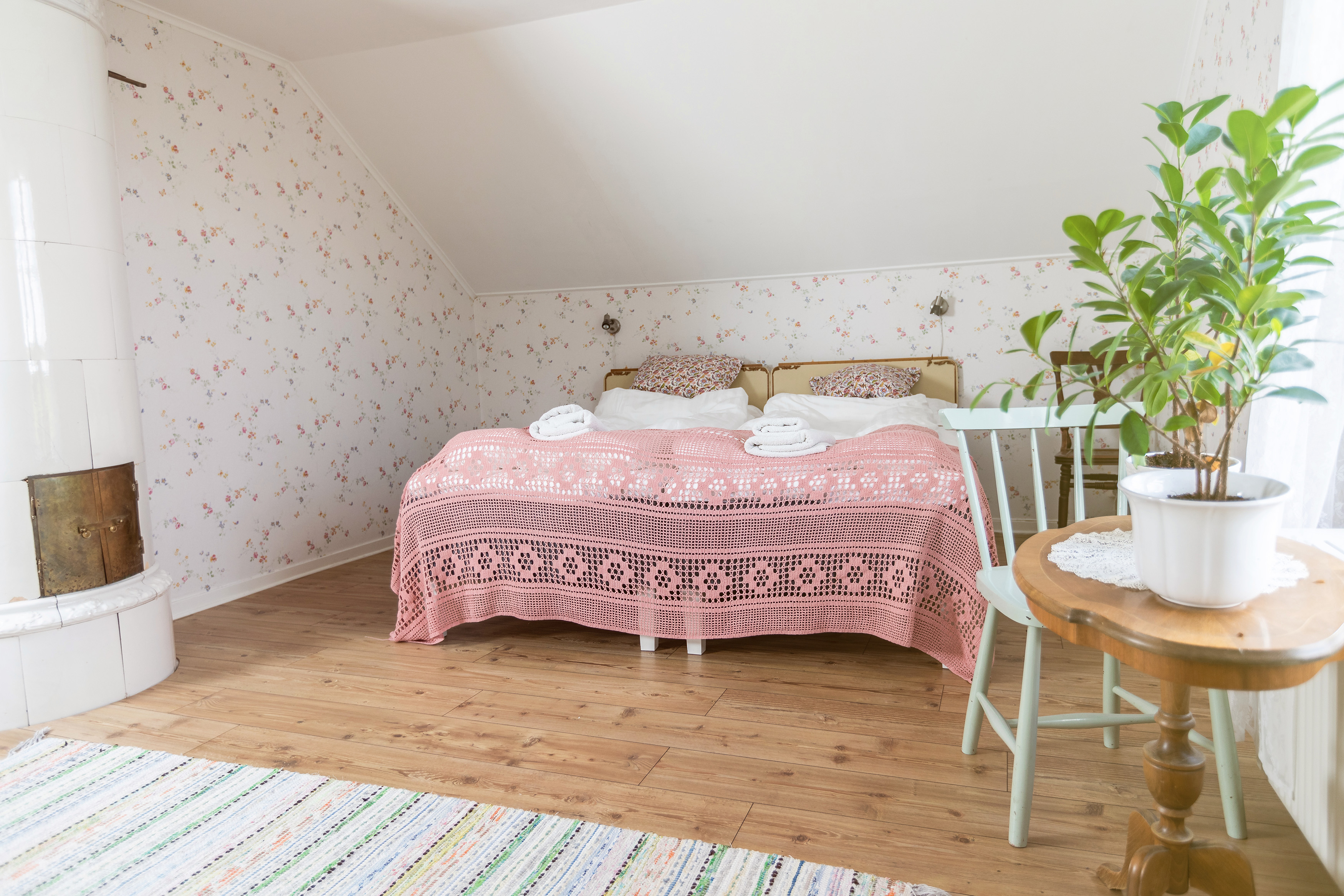 A double bed with pink crochet bedspread, wooden floor and a tiled stove.