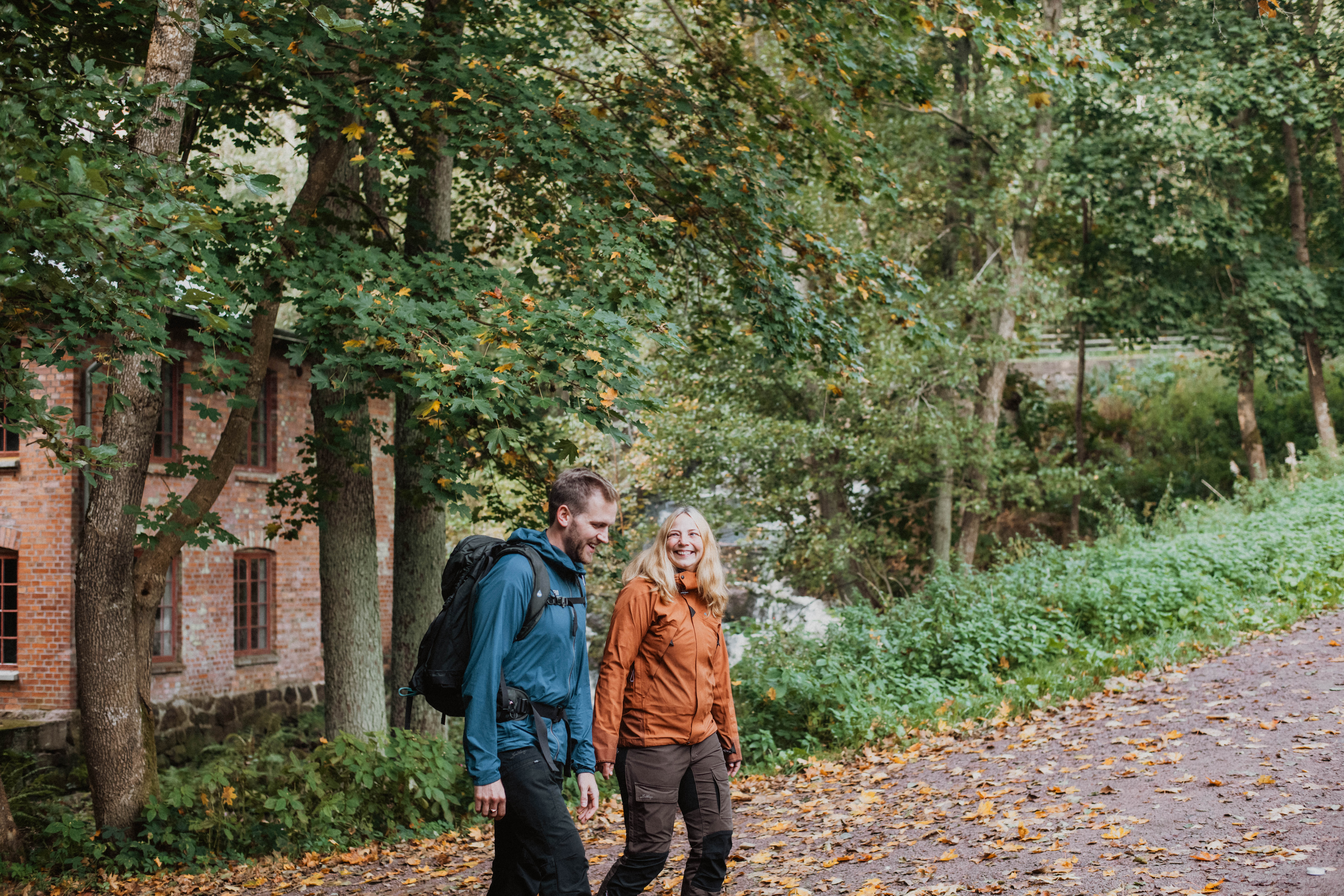 A guy with a blue jacket and a woman in an orange jacket walk on a path by an old brick building.