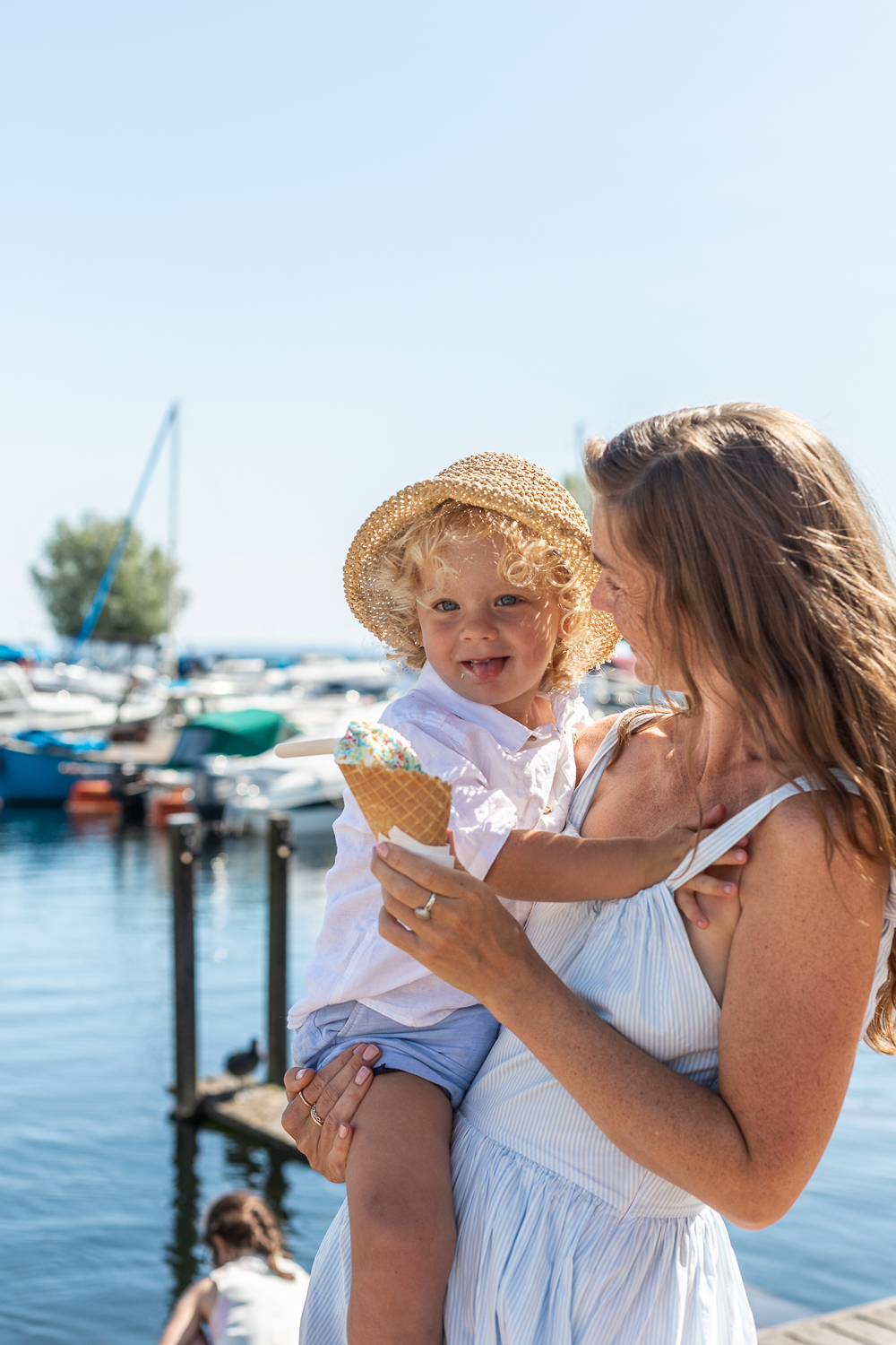 A woman is standing, holding a child in one arm and an ice cream in the other. In the background, you can see a harbor with many boats.