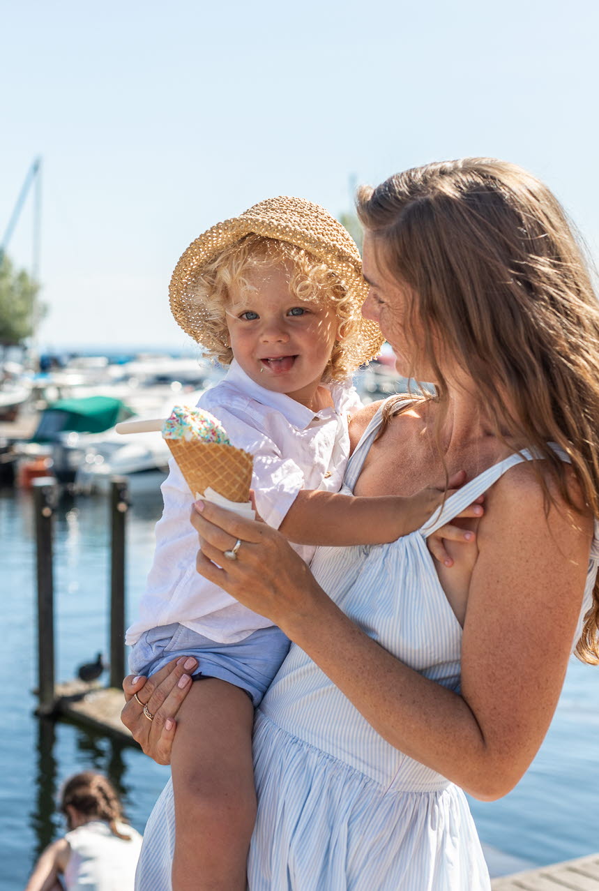 A woman is standing, holding a child in one arm and an ice cream in the other. In the background, you can see a harbor with many boats.