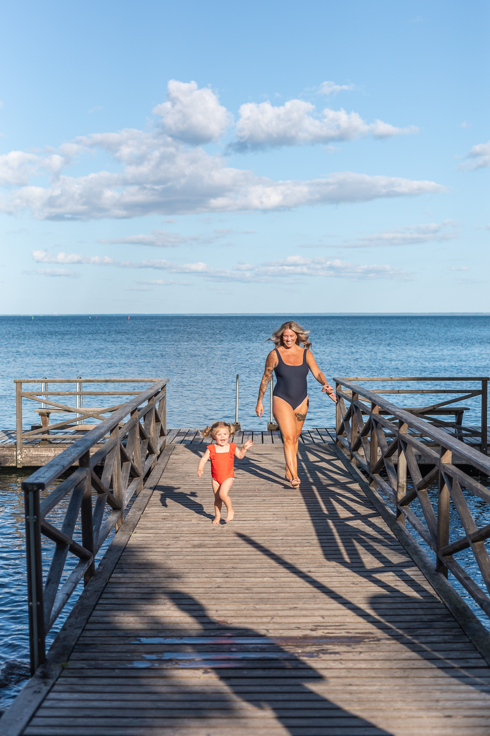A woman in a swimsuit is walking on a dock, and her daughter is running ahead of her.