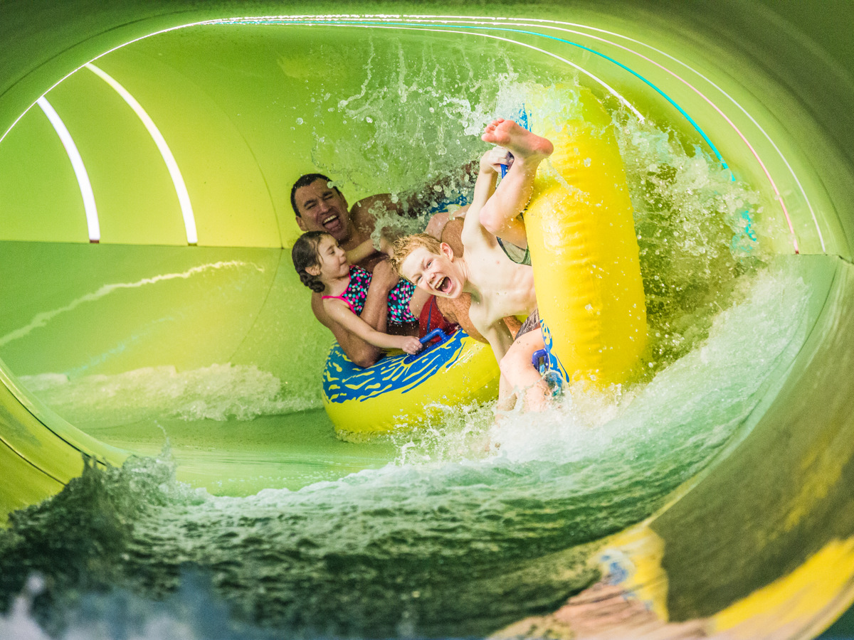 A man, a girl and a boy sit in large bath toys sliding down at full speed in a water slide