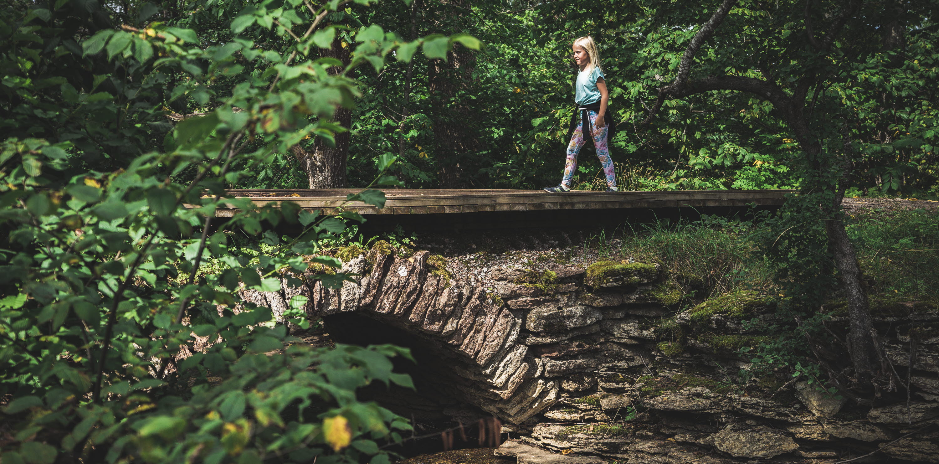 A child dressed in light clothes is walking over an arched stone bridge. The trees around the bridge are green.