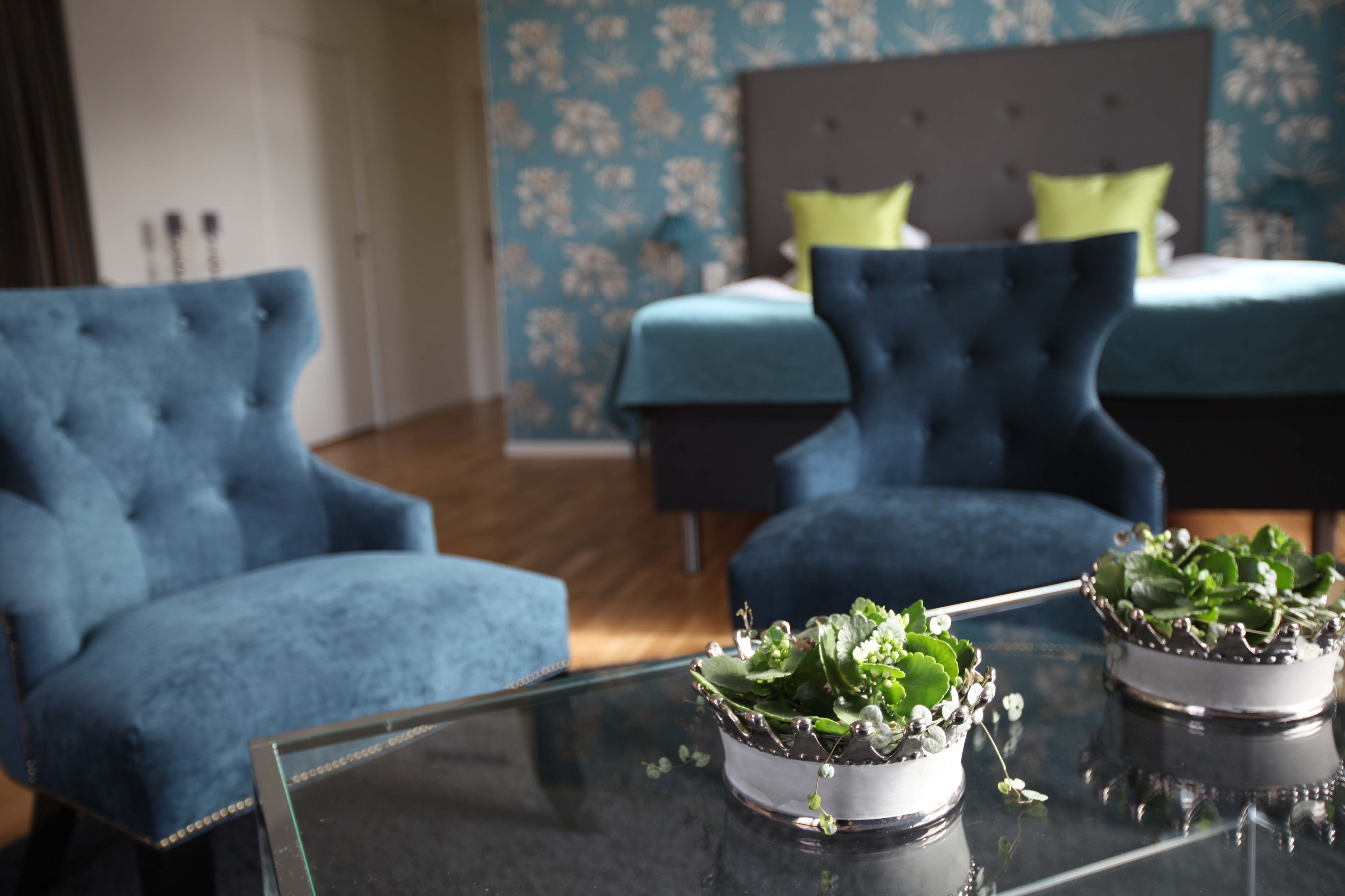 Hotelroom with double bed in the back and two blue armchairs in front. In the foreground is a table with two flowerpots.