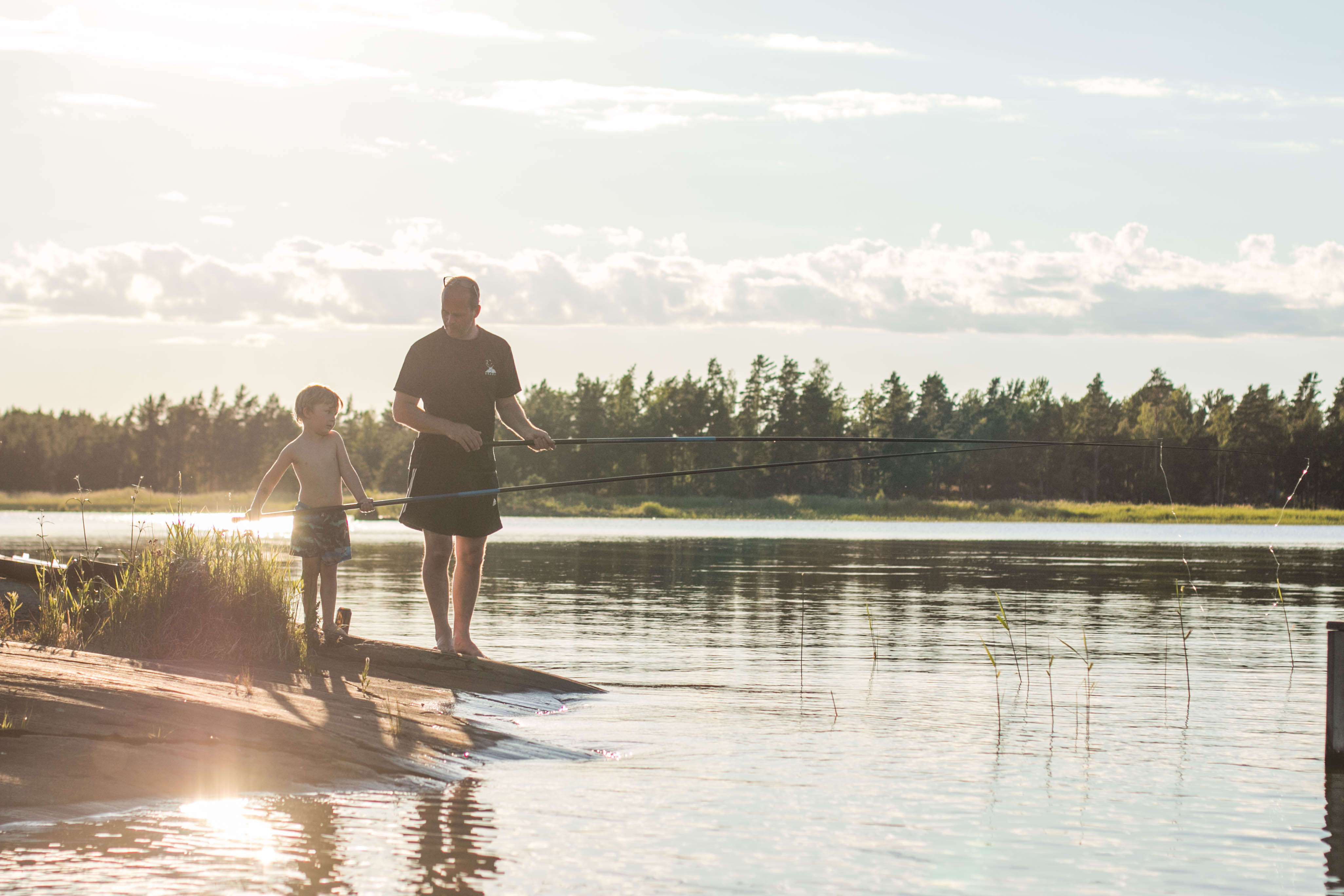 An adult and a child stand with their fishing rods in their hands by the water. Behind them you can see forest and a sunset.
