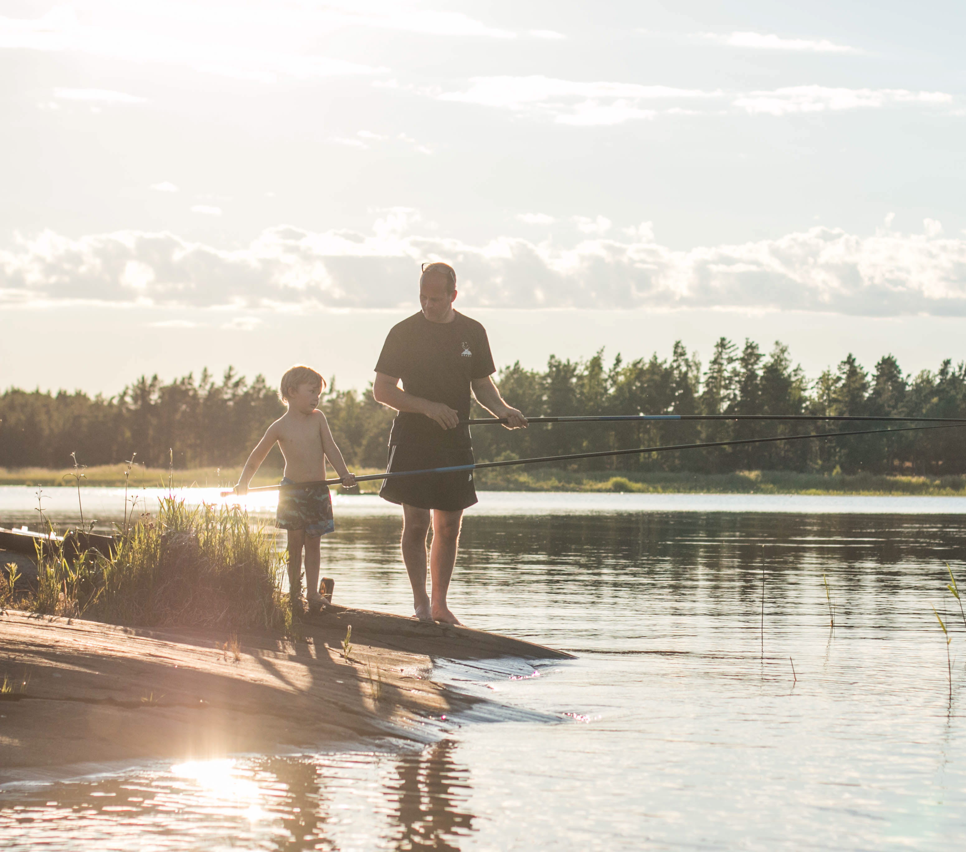 An adult and a child stand with their fishing rods in their hands by the water. Behind them you can see forest and a sunset.