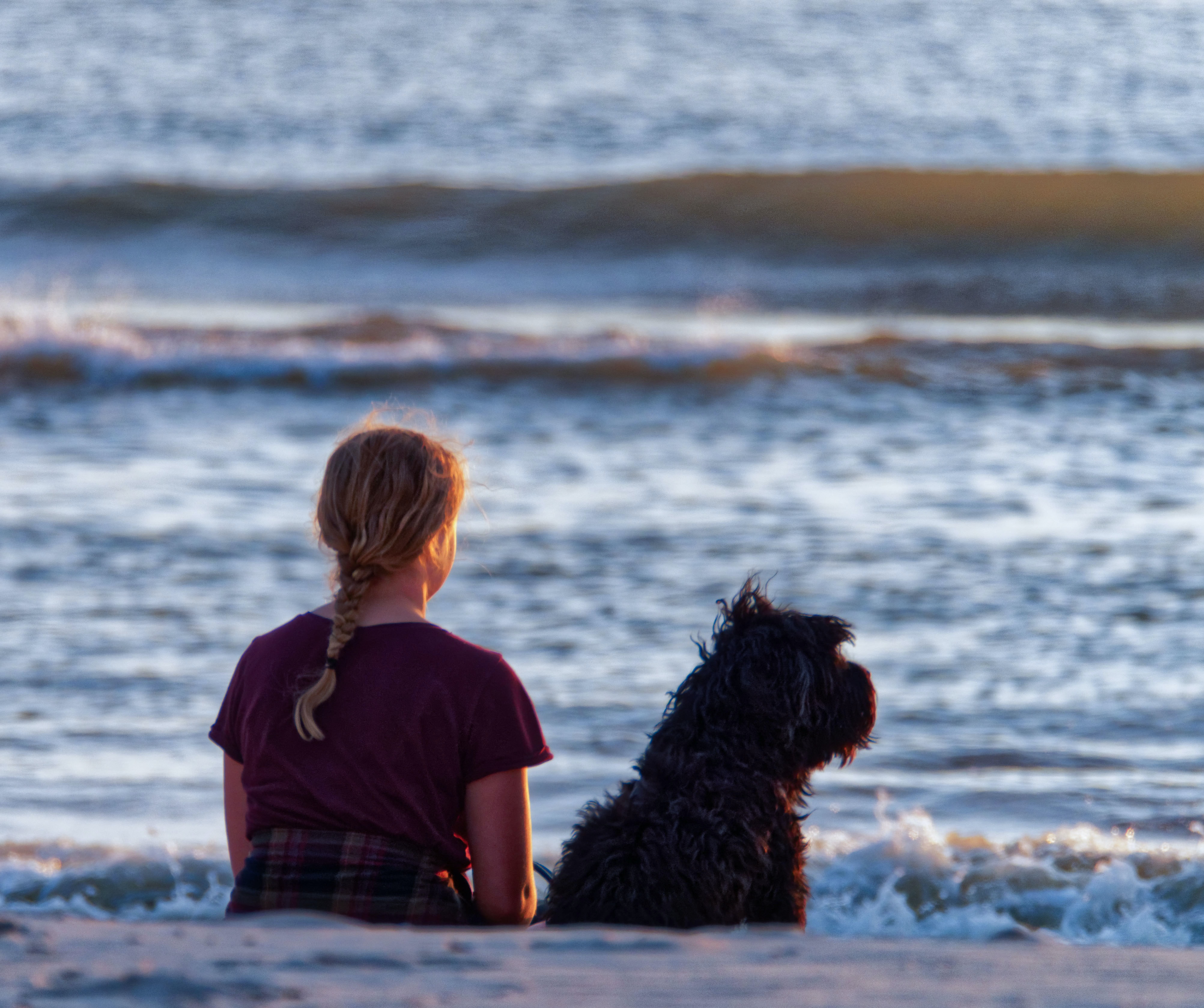 A girl is sitting on a beach with a black dog and looking out over the water.

