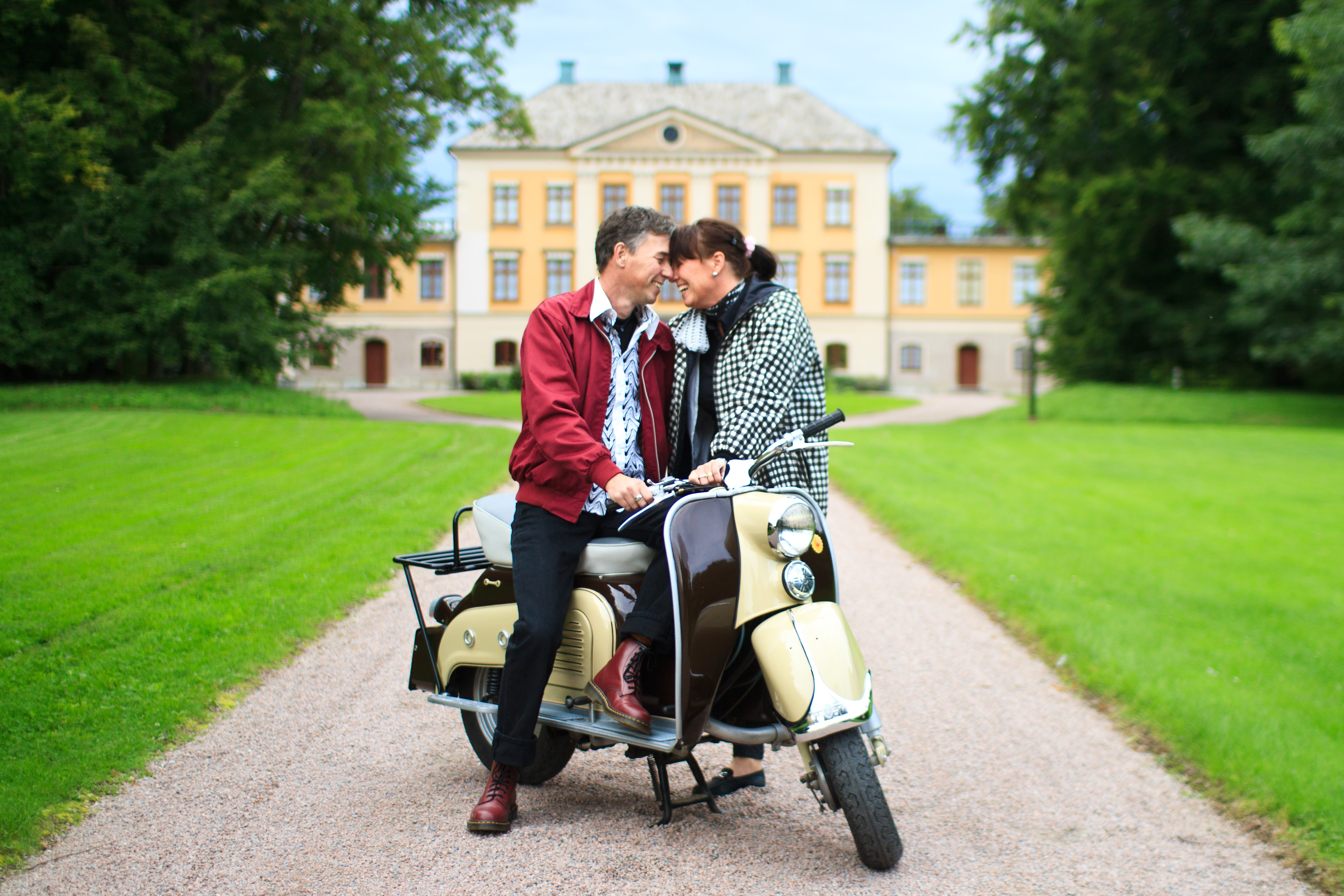 Couple on scooter in front of a yellow mansion.
