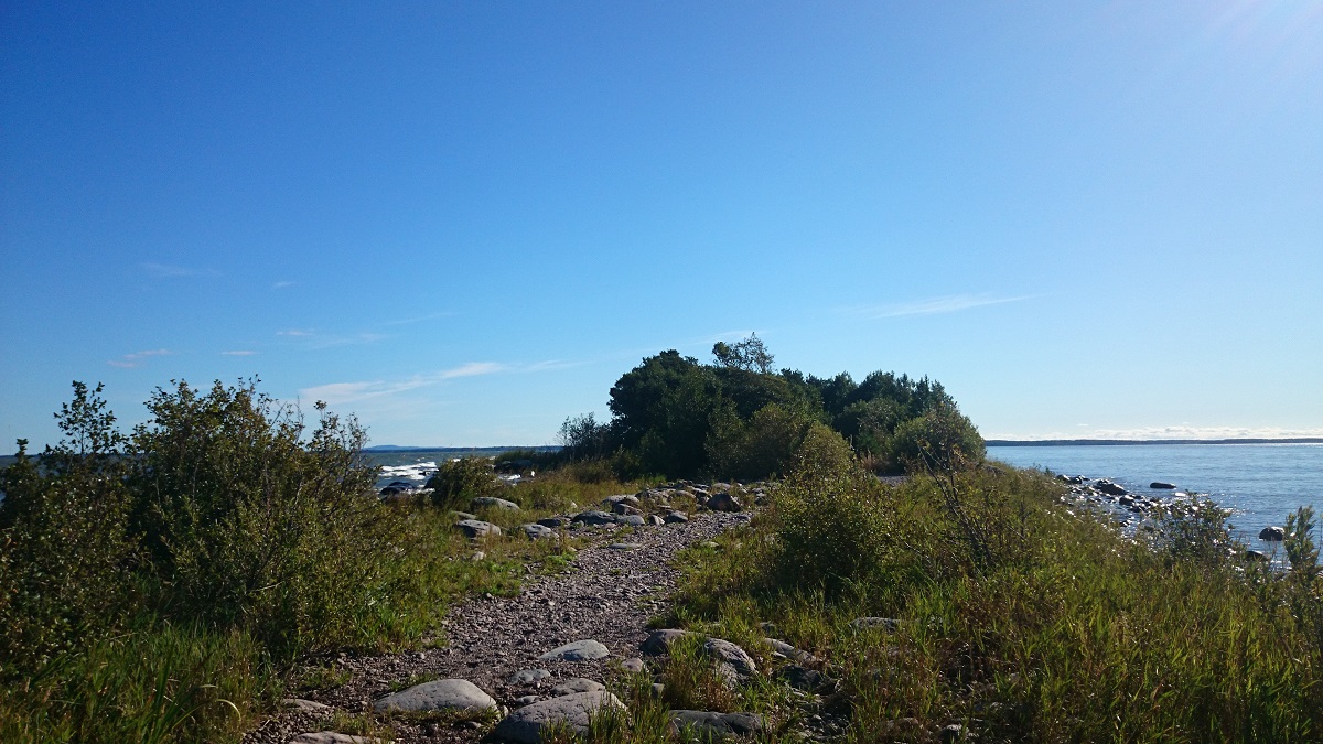 Gravel path with grass on the side. In the background you can see water and clearblue sky.