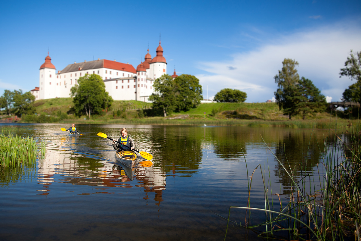 Two people kayak on still water next to land on a sunny summer day. In the background is a large white pompous Baroque castle.