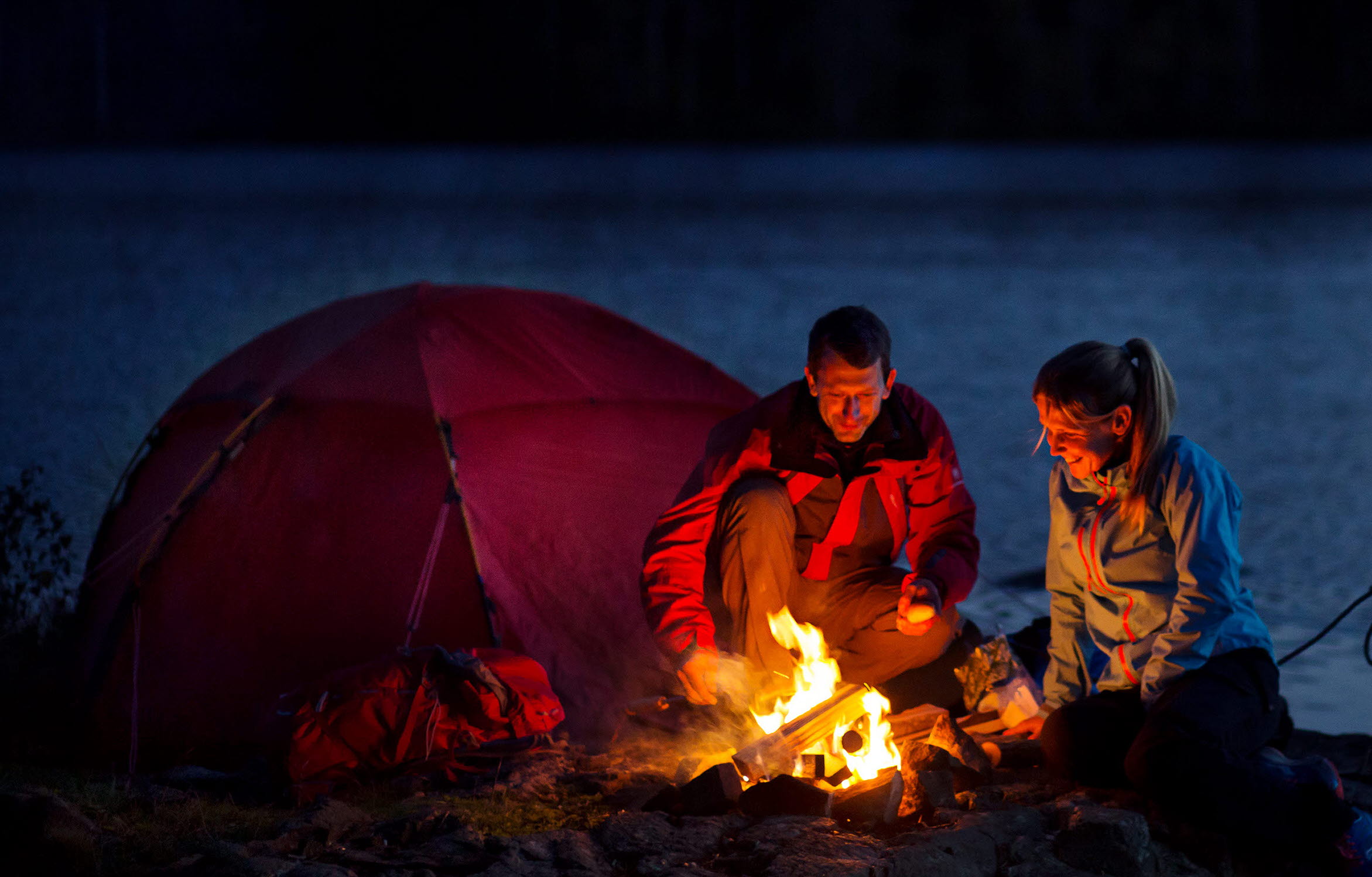 A man and a woman sitting by a bonfire outside their tent at night. Behind them there is a lake and a forest. Beside them there are a canoe.