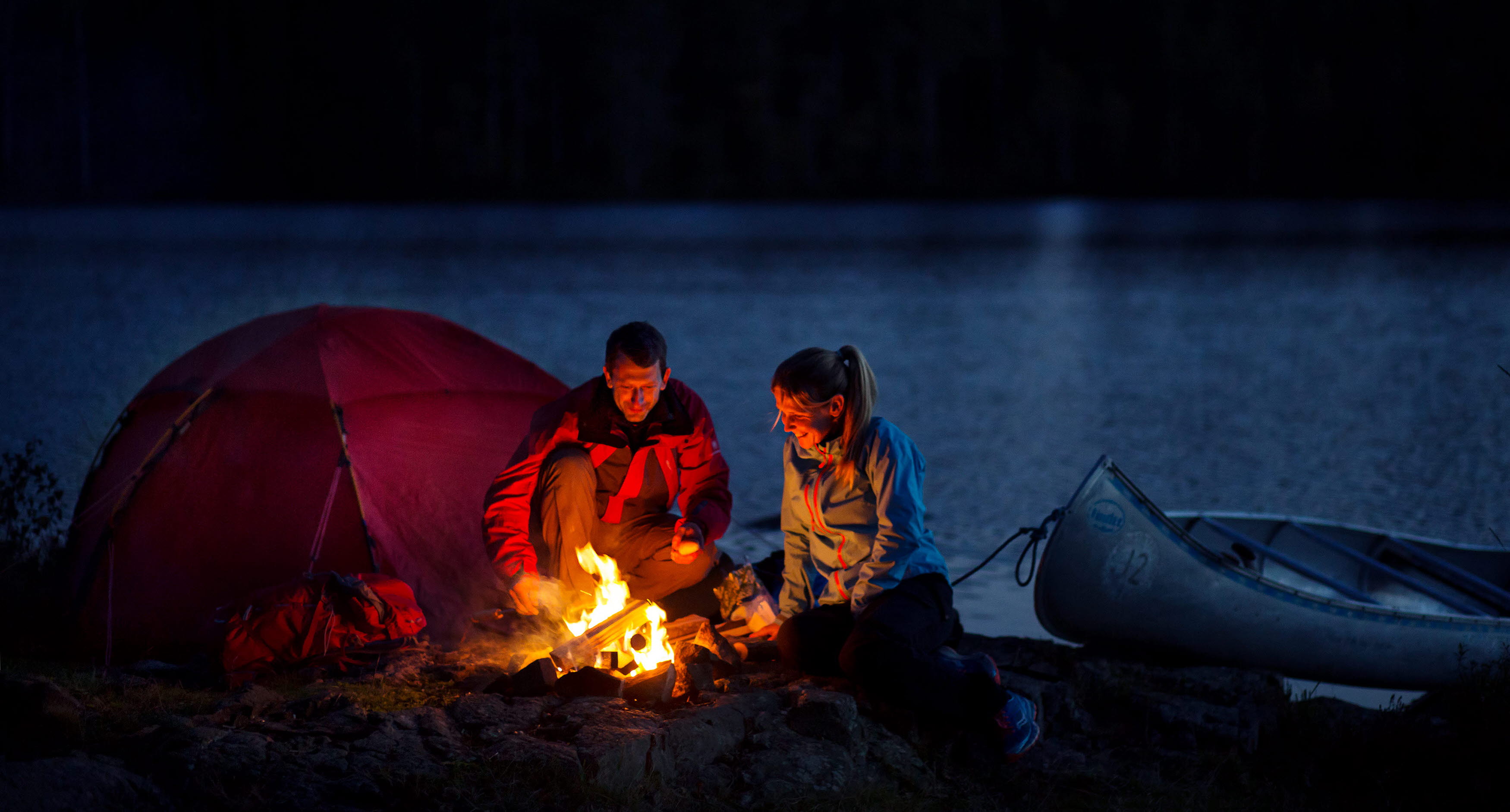 A man and a woman sitting by a bonfire outside their tent at night. Behind them there is a lake and a forest. Beside them there are a canoe.