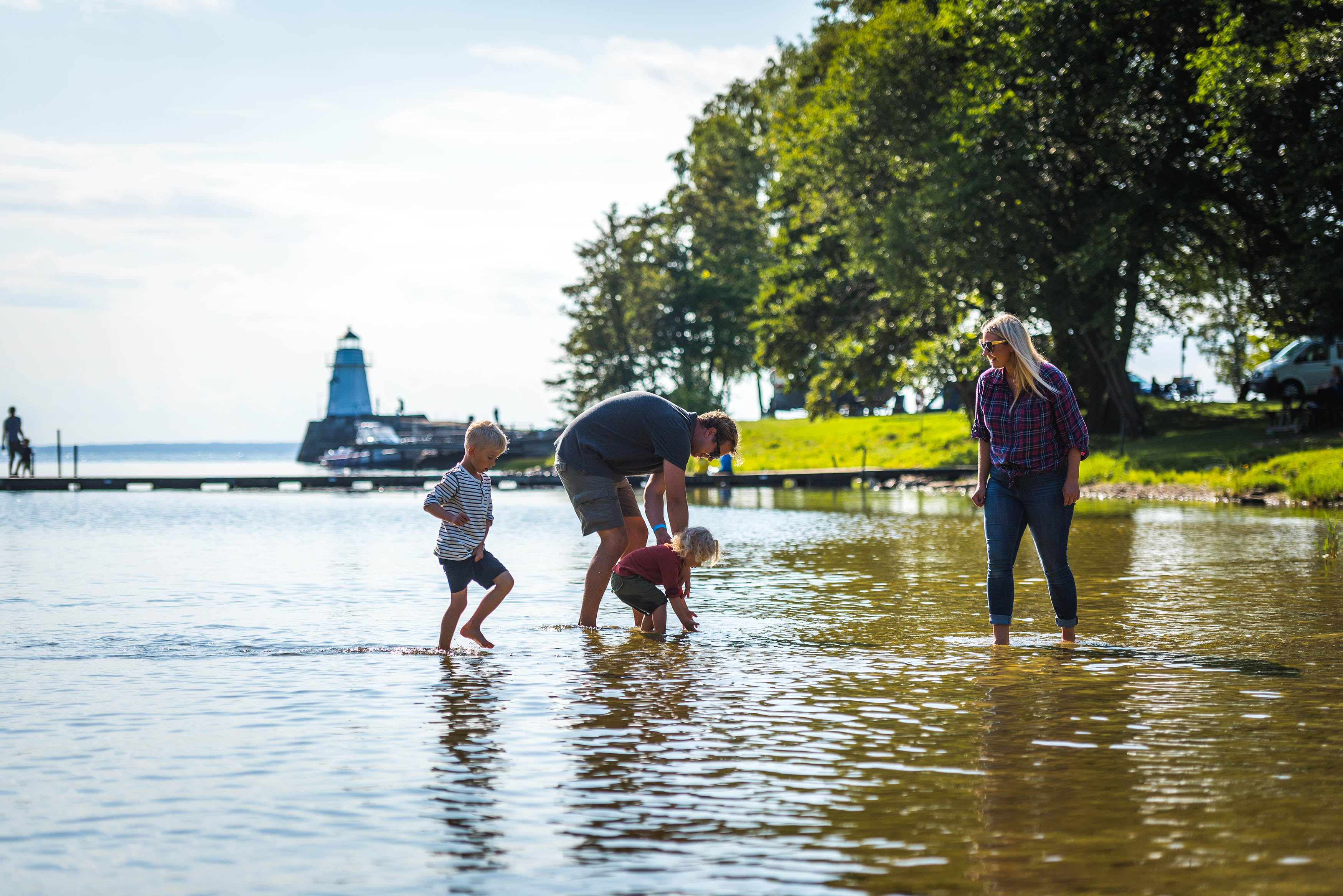 A family is wading in the water. The sun is shining and in the background you can se some trees an an old light house.