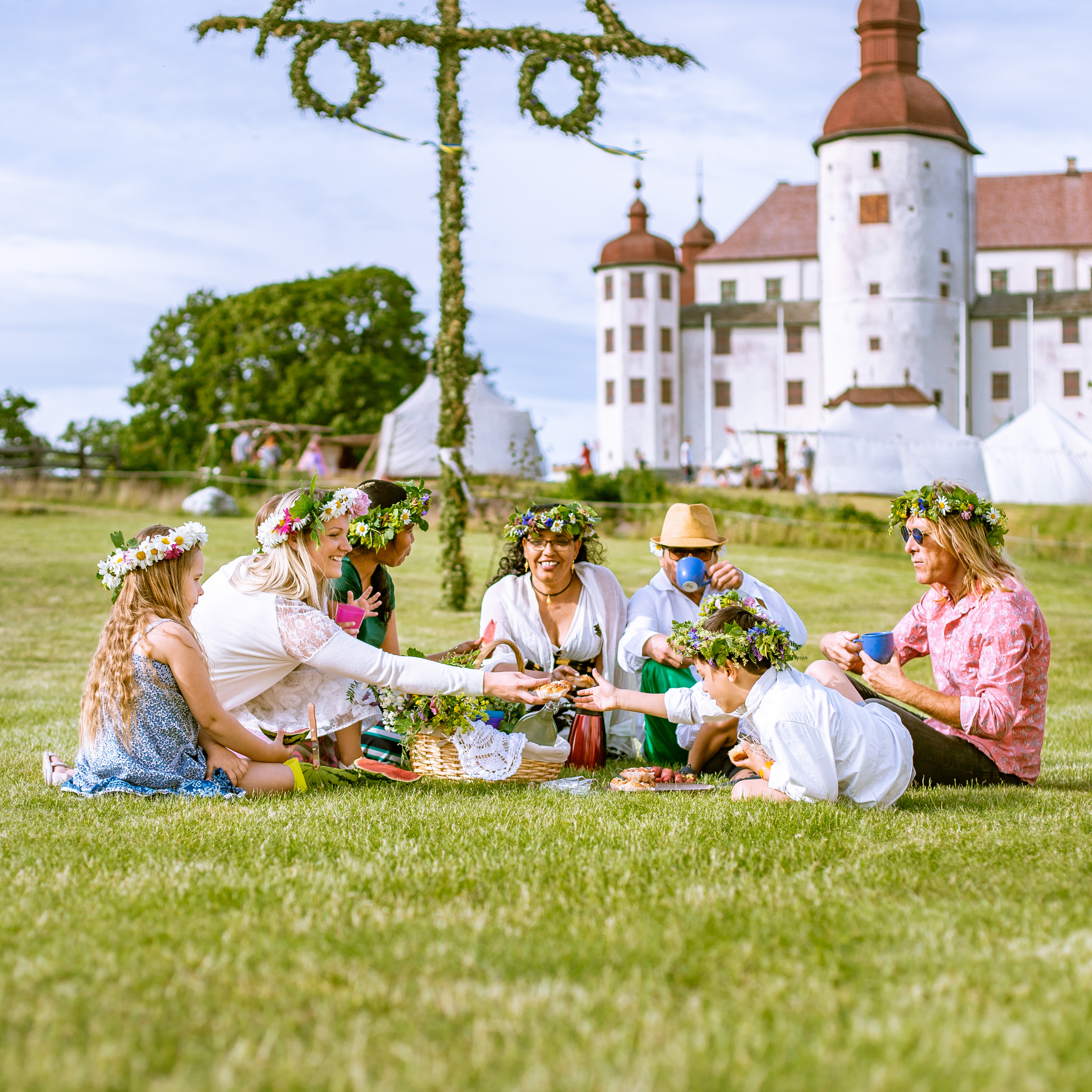 Midsummer on Läckö. In the picture there is 7 people sitting in a ring and having coffee. In the background, Läckö Castle and a dressed midsummer pole can be seen.
