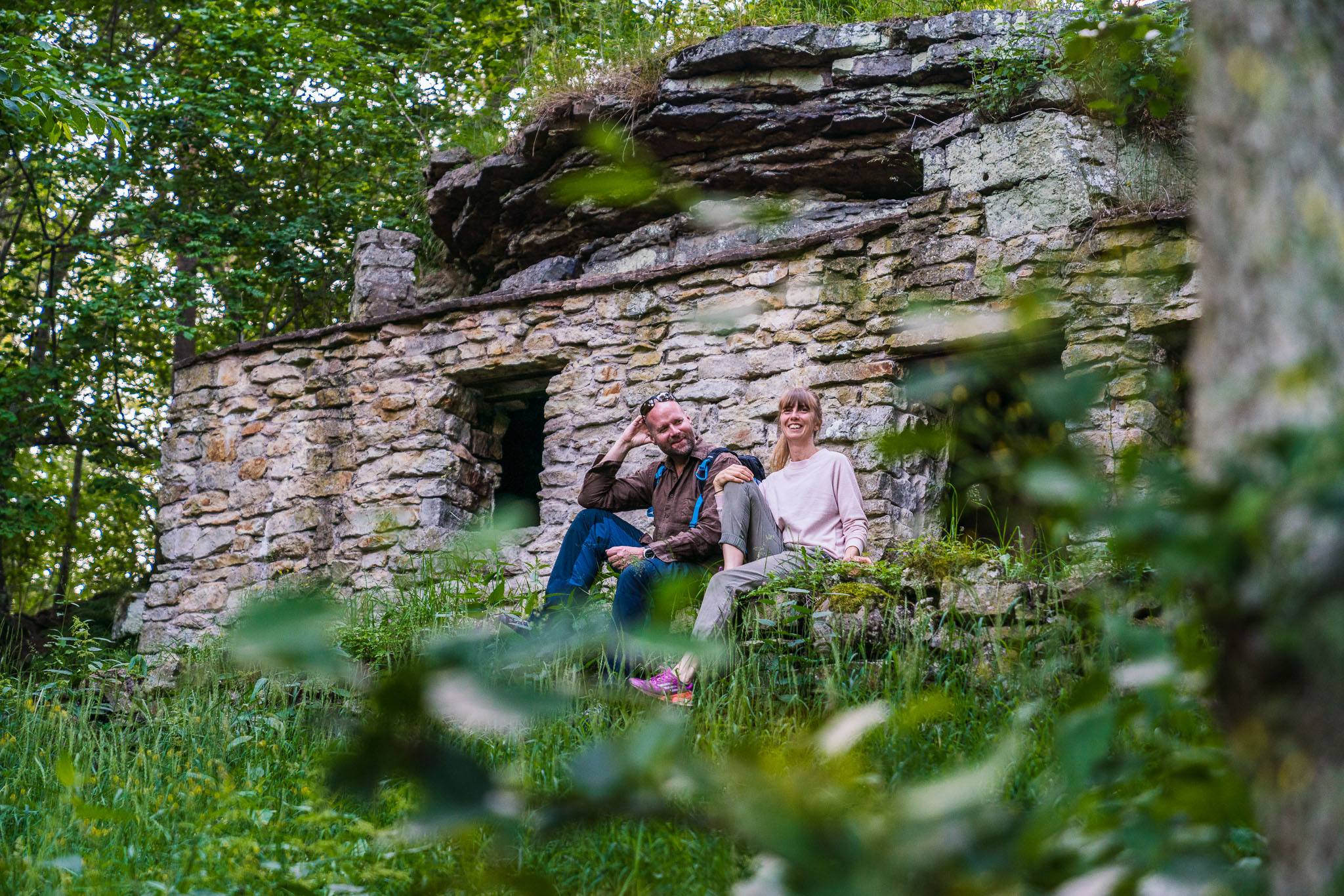 Two people, a man and a woman, sitting in front of a stone cave in the forest.
