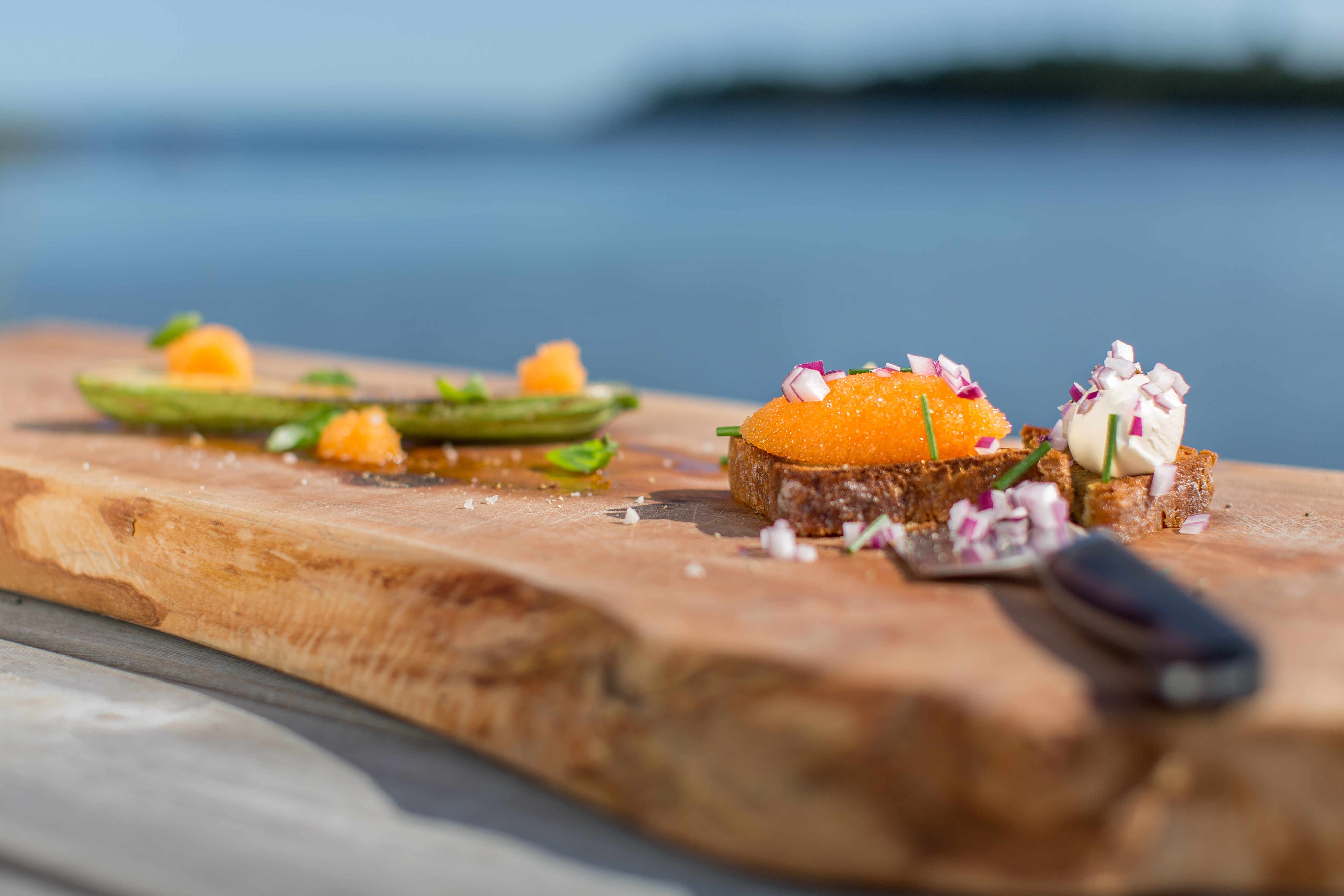 Slice of bread full of whitefish roe and chopped red onion served on a plank. In front is a knife.