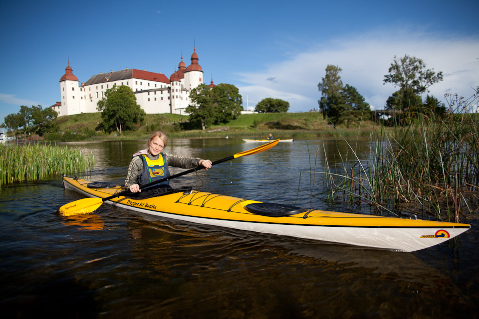 A woman sits in a kayak and looks into the camera. Behind her is the magnificent white Läckö Castle.