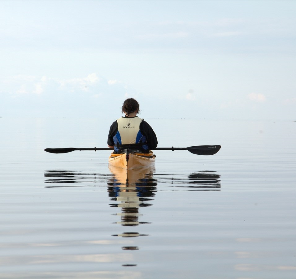 A person sits in a kayak with his back to the camera. The water is still and goes in the same gray-blue tone as the sky.