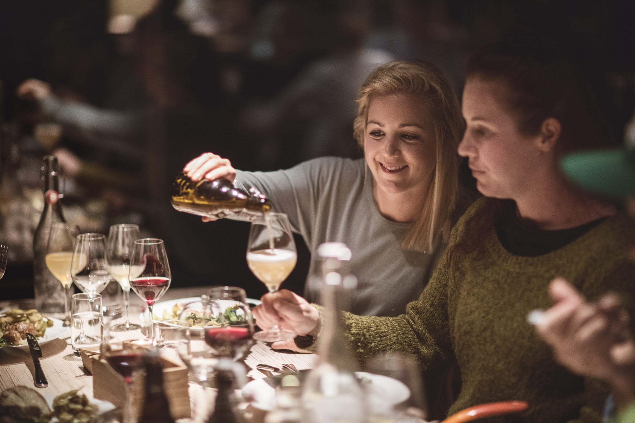 Two women having dinner. One is pouring drinks to the other one.