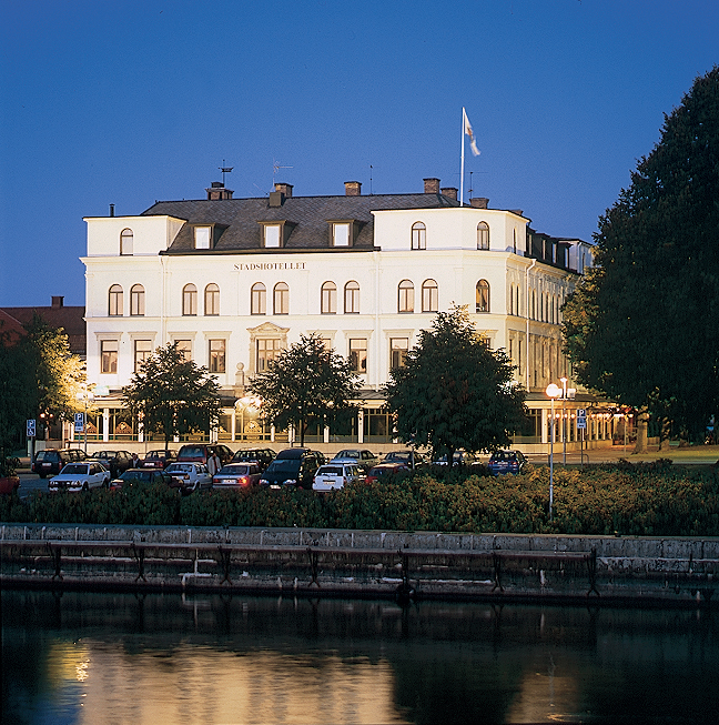 A large old white building with black roof right next to a river. It is evening and the lighting illuminates the building in a beautiful way.
