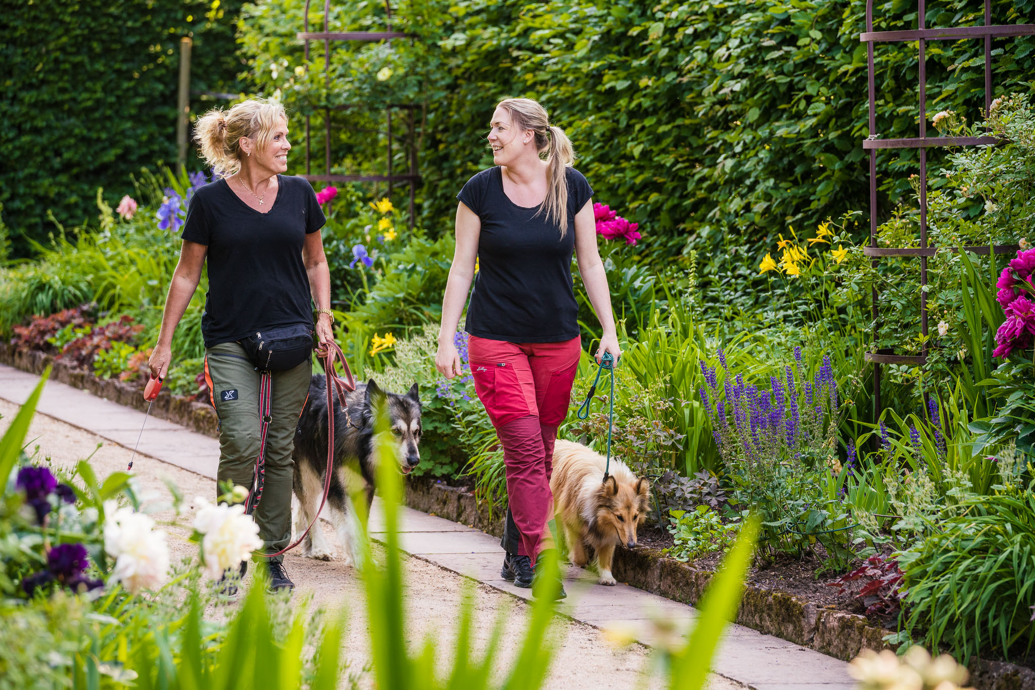 Two women with dogs dressed in hiking clothes in a beautiful garden environment.