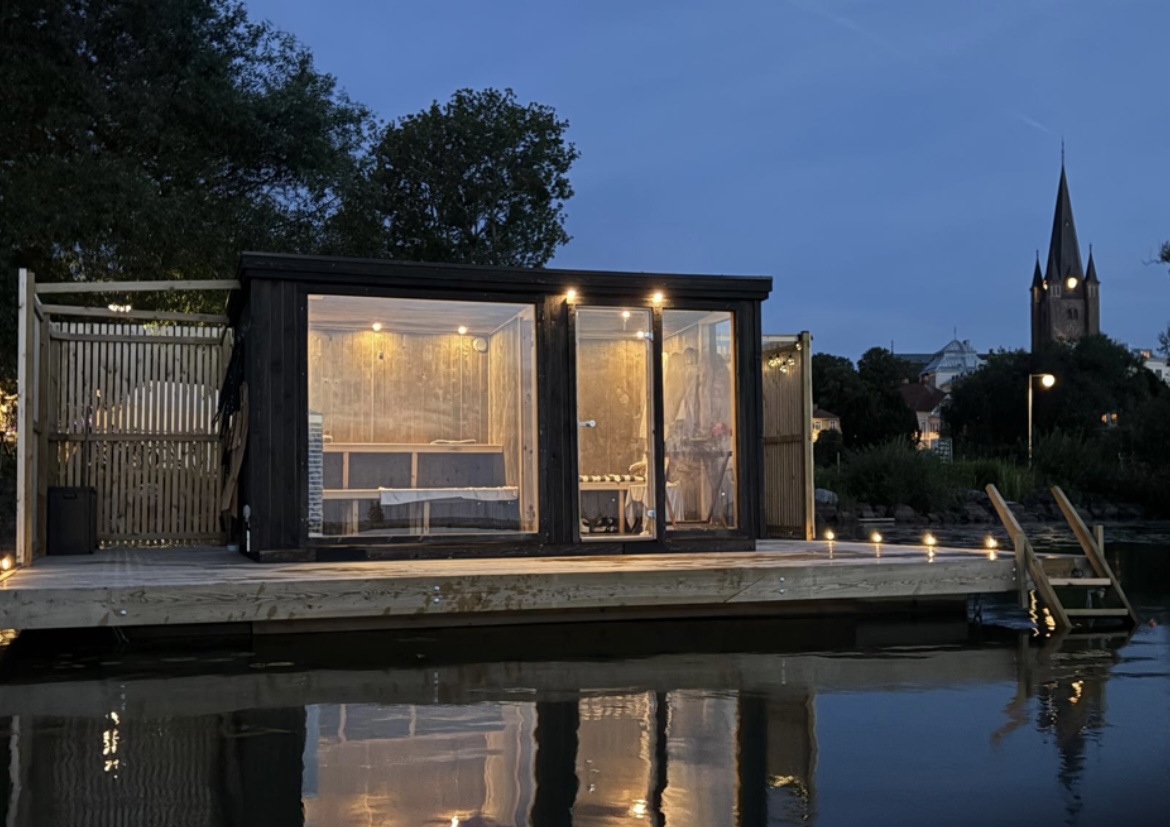 Sauna raft at dusk illuminated from the inside with a reflection in the water in front and with the cathedral in the background.