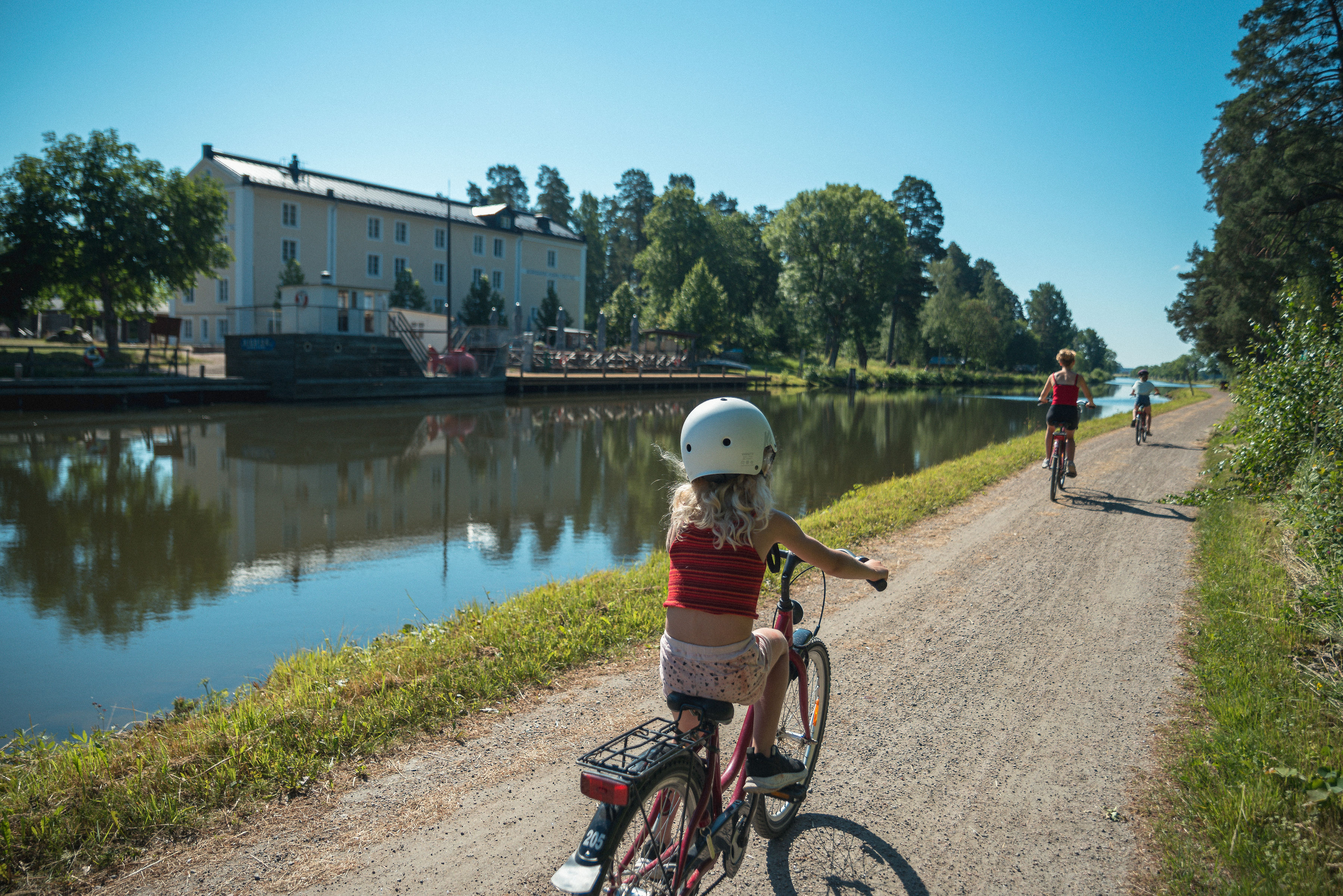 Three people cycling on a gravel road along the Göta Canal. In the foreground a child with a helmet on. On the other side of the canal Norrkvarn's large yellow four-storey hotel building.