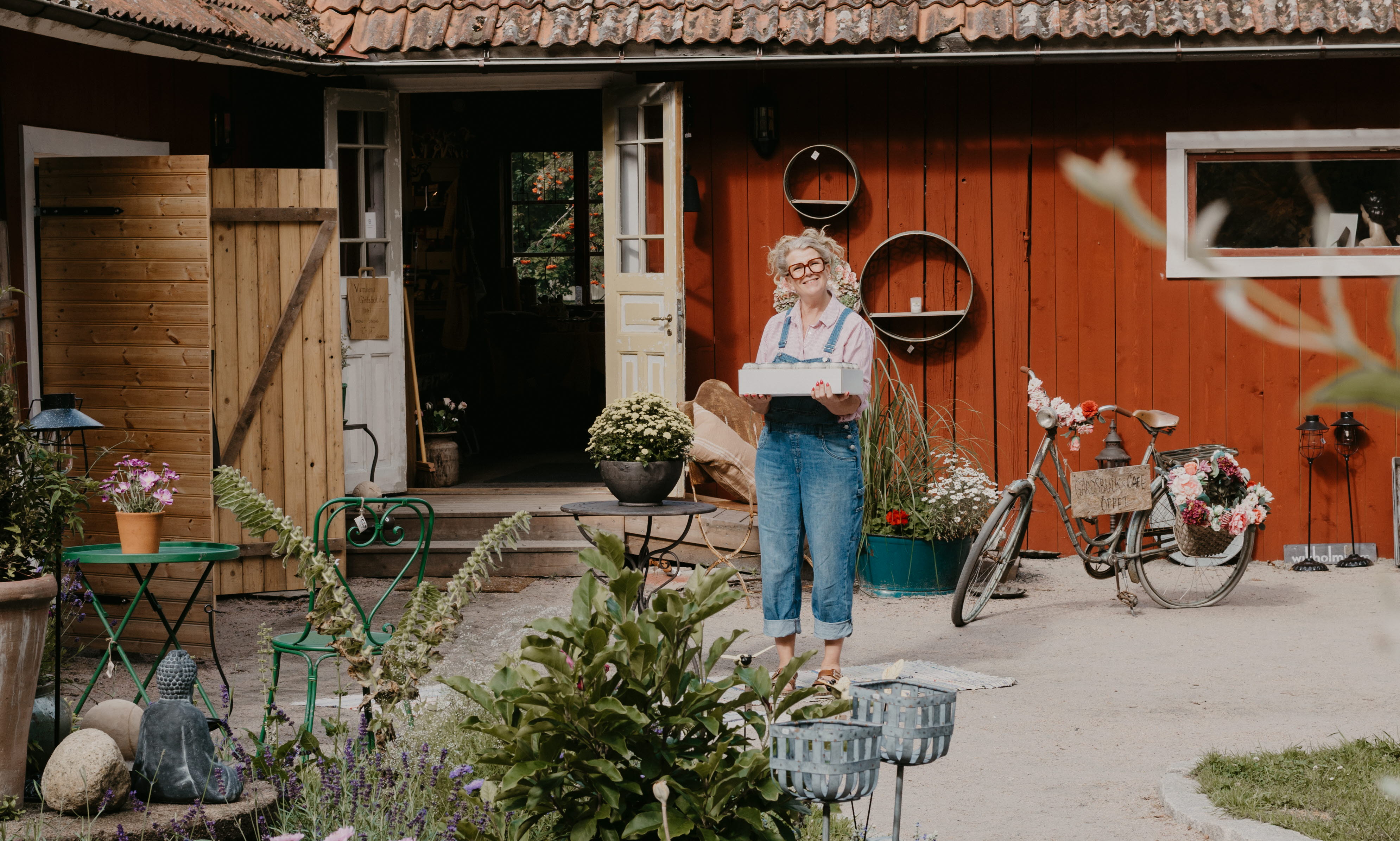 A woman is holding a box. She is wearing blue suspenders and is standing in a courtyard. In the foreground you can see a nice planting.
