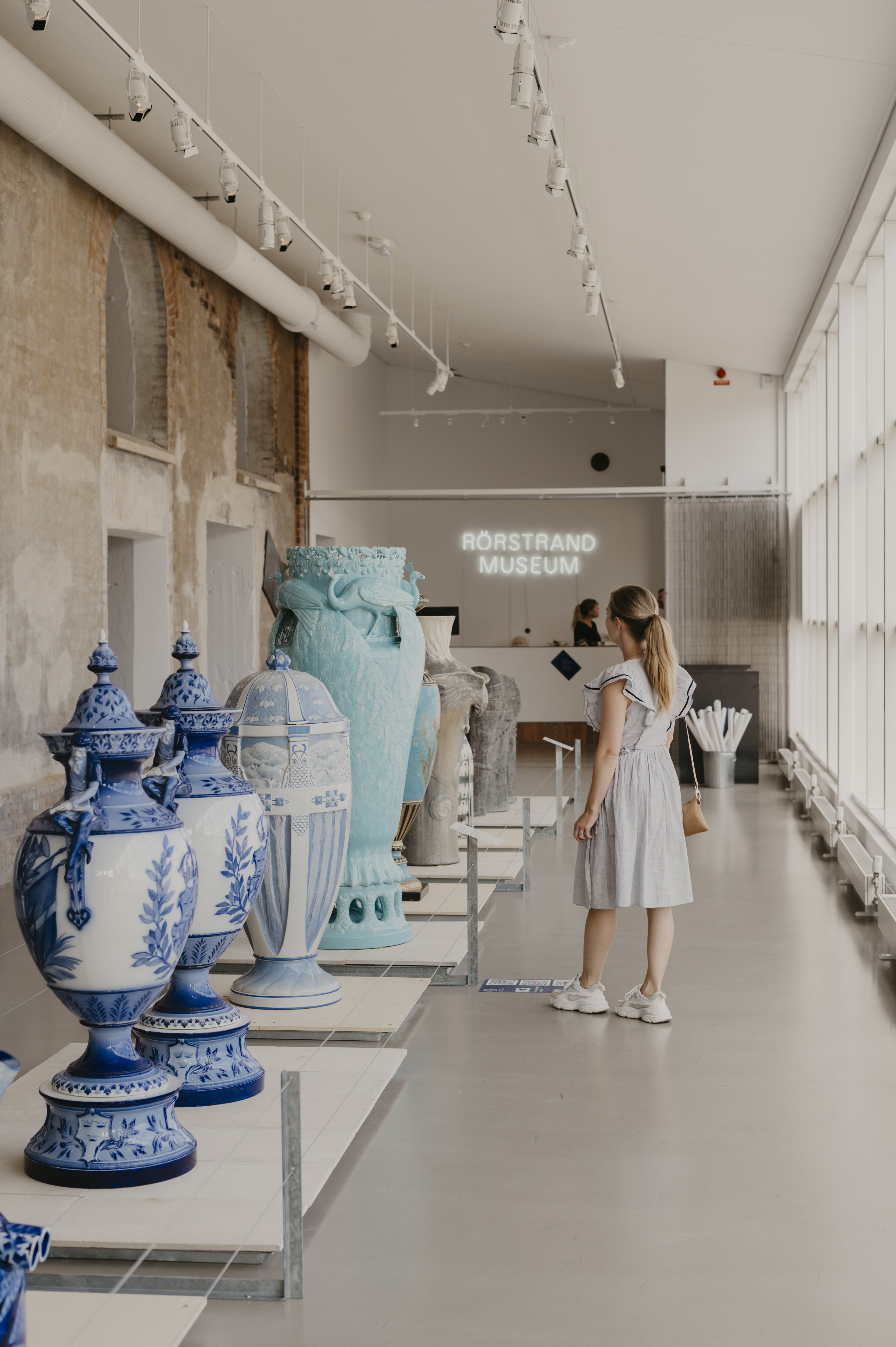 A woman in a bright dress walks around inside a museum where large porcelain pieces are lined up along one wall.
