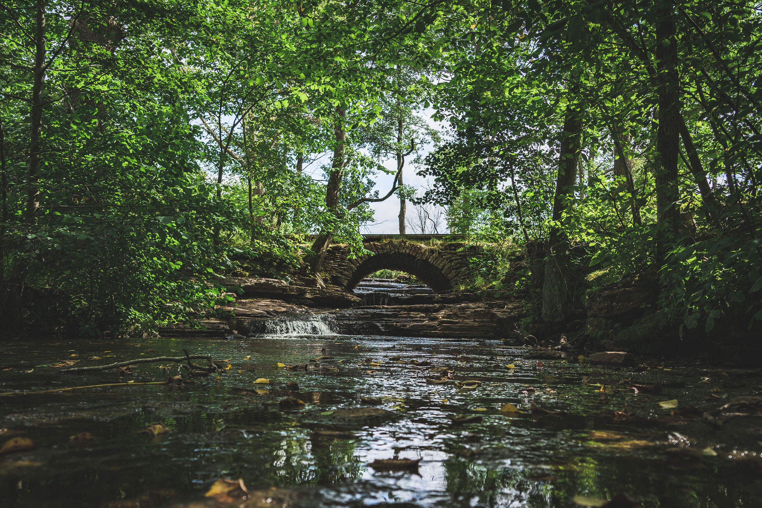 A quiet waterfall flowing under a beautiful old stone bridge with greenery all around.
